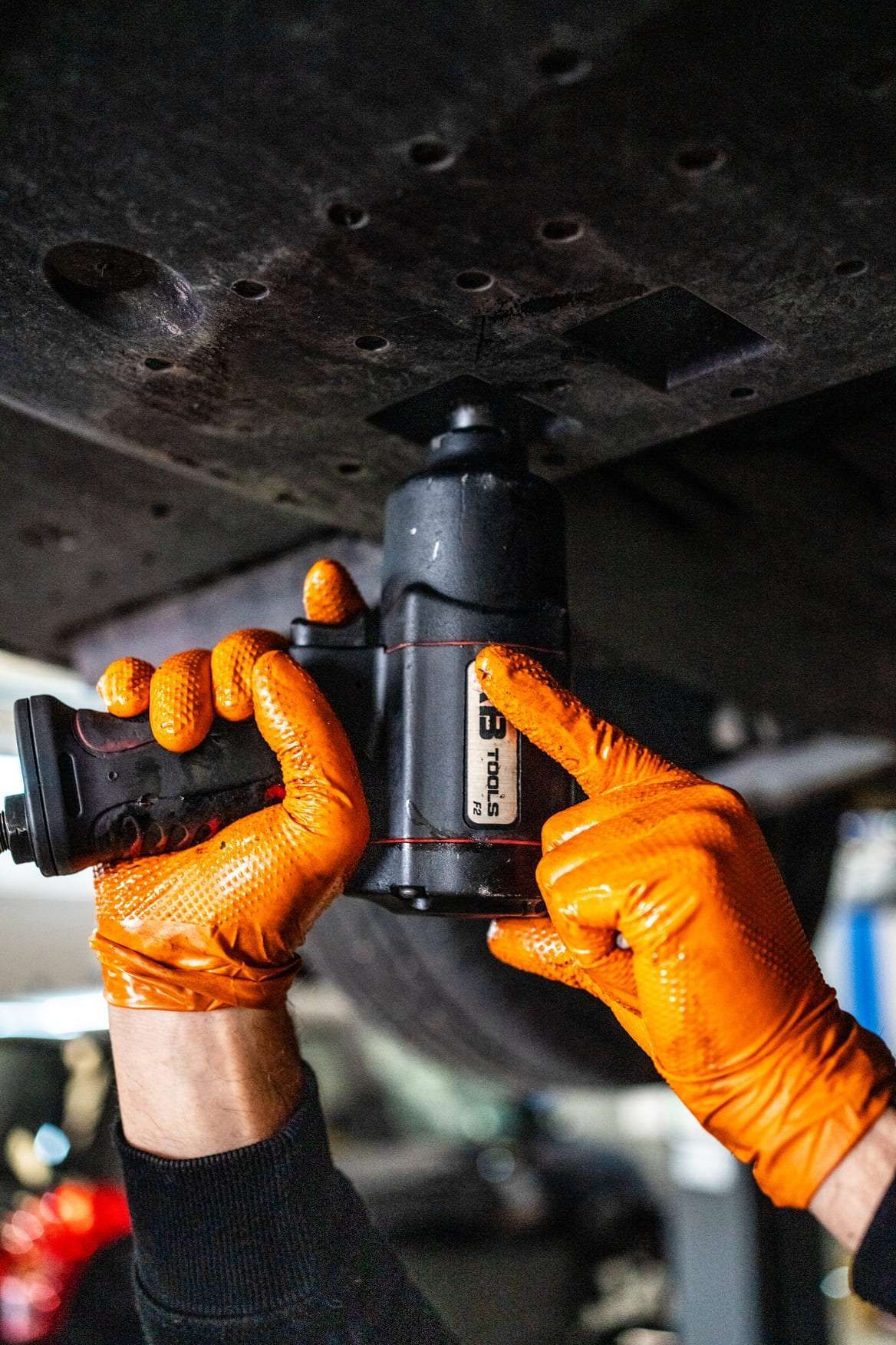 Una persona que lleva guantes de color naranja sostiene un gato debajo de un coche.