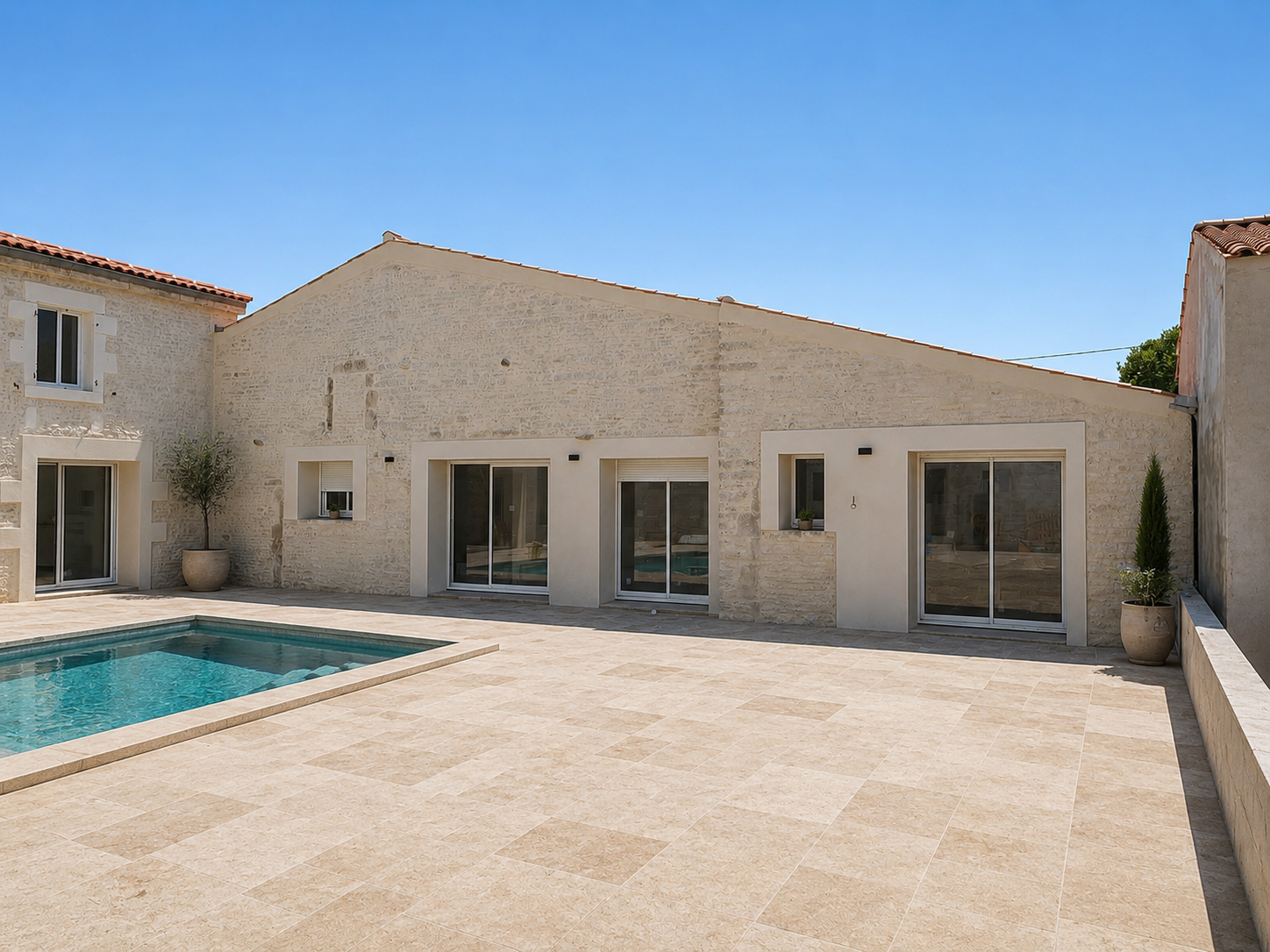 Terrasse en pierre et piscine turquoise jouxtant une villa couleur crème avec de grandes baies vitrées coulissantes sous un ciel d'un bleu limpide.