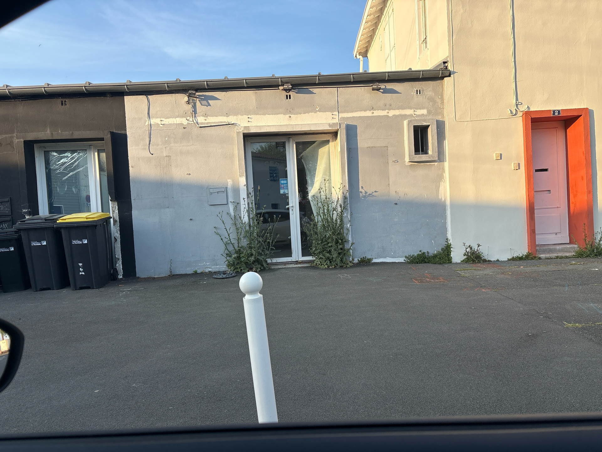 Extérieur d'un bâtiment beige avec une porte bleue, une porte latérale orange, des poubelles et un parking pavé.