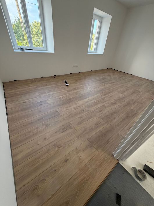 Pièce vide avec parquet clair, murs blancs, deux fenêtres et une porte visible.