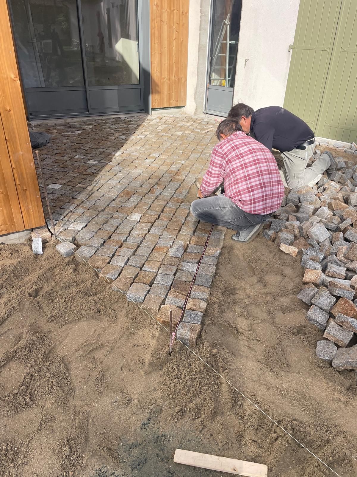 Deux ouvriers posent des pavés sur une allée sablonneuse à côté d'un bâtiment, avec des tas de pierres à proximité.