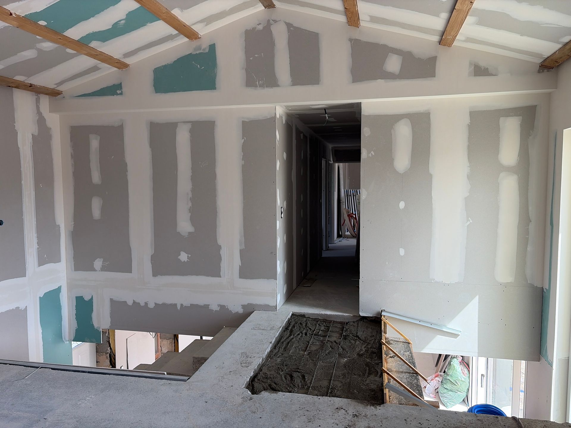 Intérieur d'une pièce inachevée avec cloisons sèches, poutres apparentes et une grande ouverture dans le sol donnant sur un couloir.