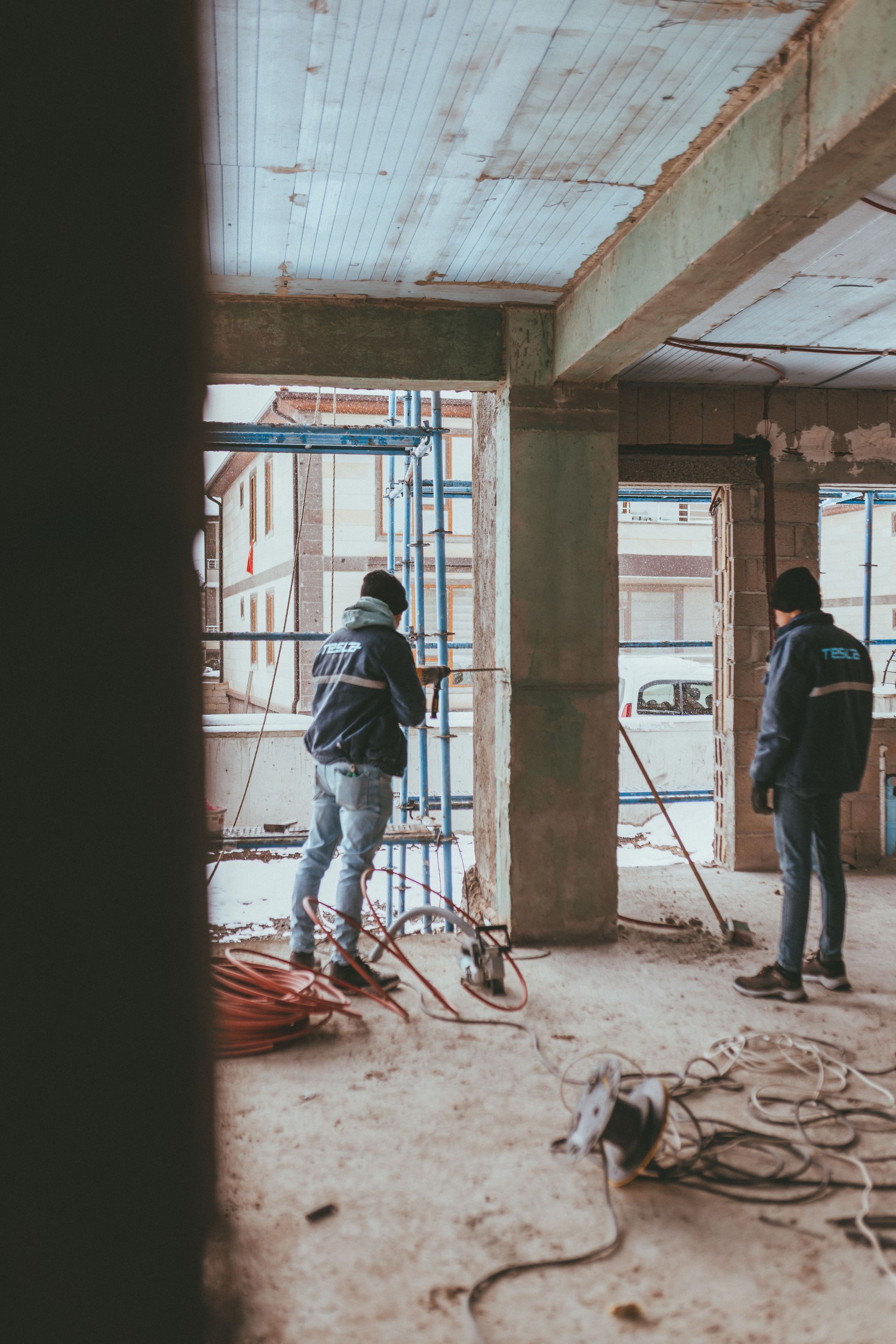 Deux ouvriers dans un bâtiment poussiéreux et inachevé, avec des échafaudages et des colonnes de béton apparentes.