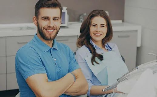Un hombre y una mujer están sentados en una silla dental y sonríen a la cámara.