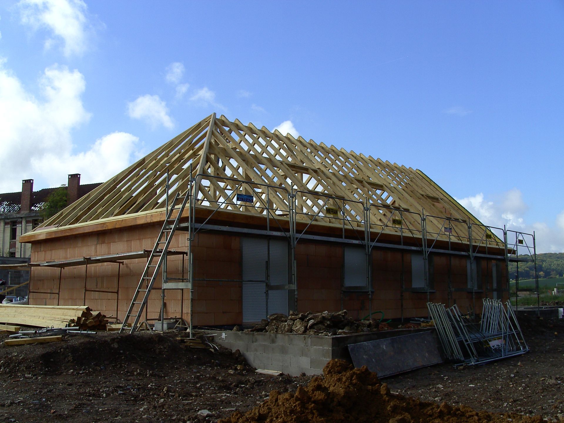 Bâtiment en construction : charpente de toit en bois sur une structure en briques, échelle appuyée contre le mur, ciel bleu.
