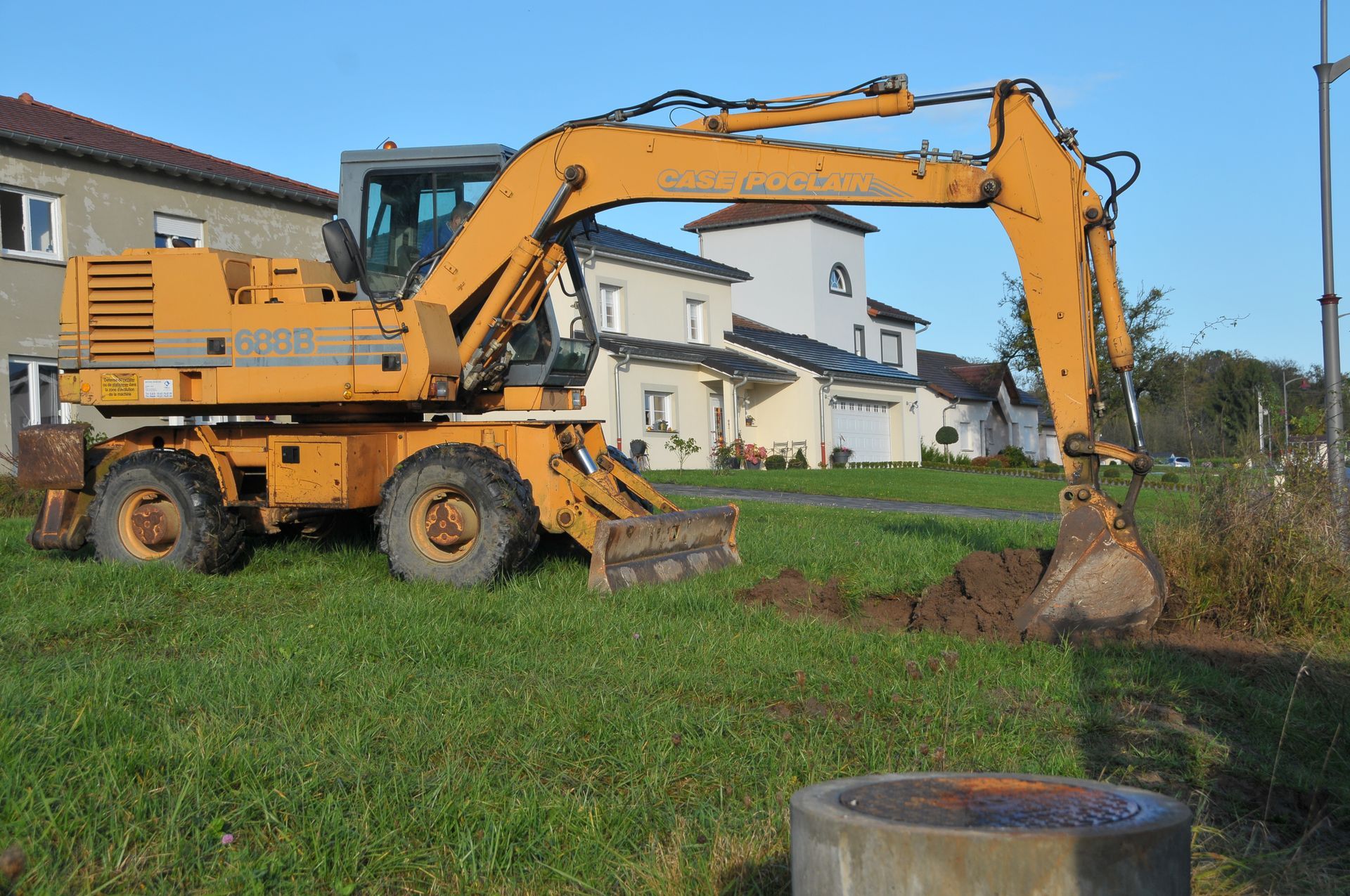 Une pelleteuse jaune creuse une tranchée dans une zone rurale herbeuse. Des montagnes et des arbres sont visibles en arrière-plan.