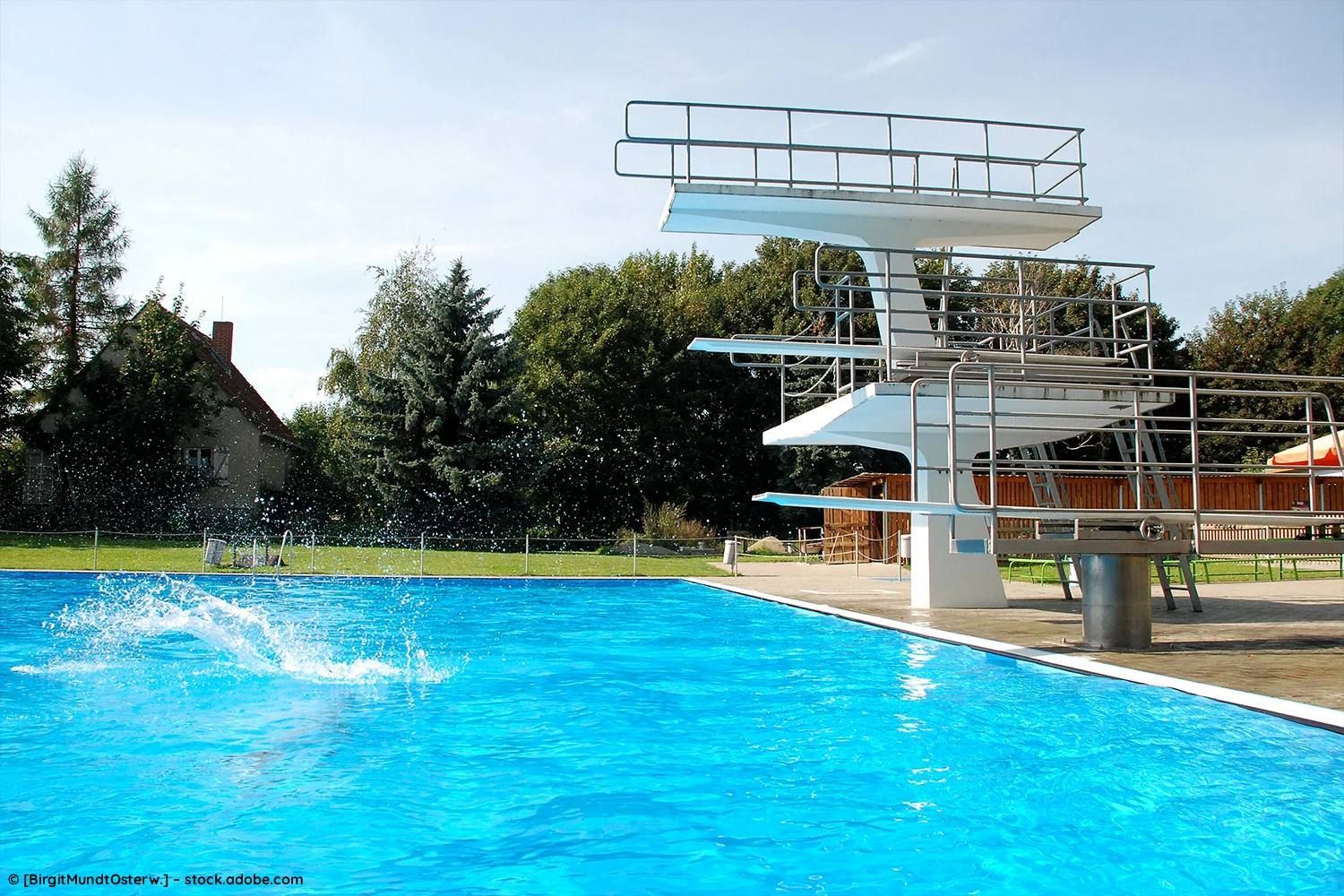 Freibad mit mehrstufigem Sprungturm und spritzendem Wasser im Vordergrund an einem sonnigen Tag.