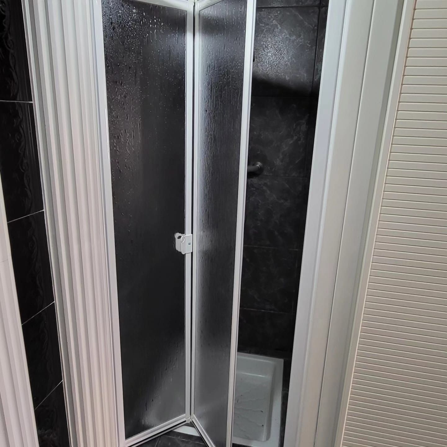 Una puerta de ducha plegable de vidrio con marco blanco está ligeramente abierta y muestra el interior de la ducha con azulejos oscuros.