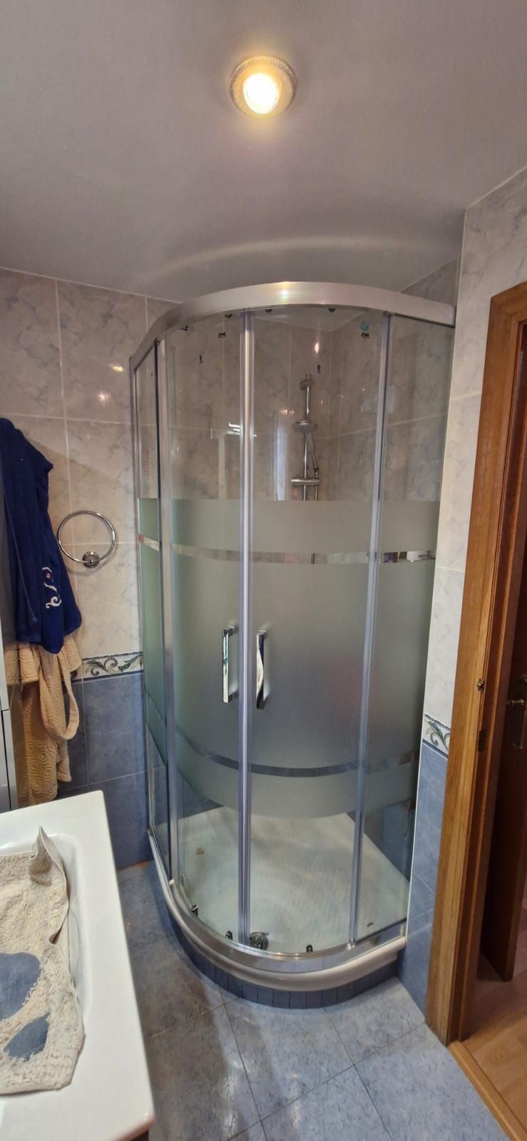 Una cabina de ducha en un baño con puertas de cristal. Las paredes están alicatadas y se ve una puerta.