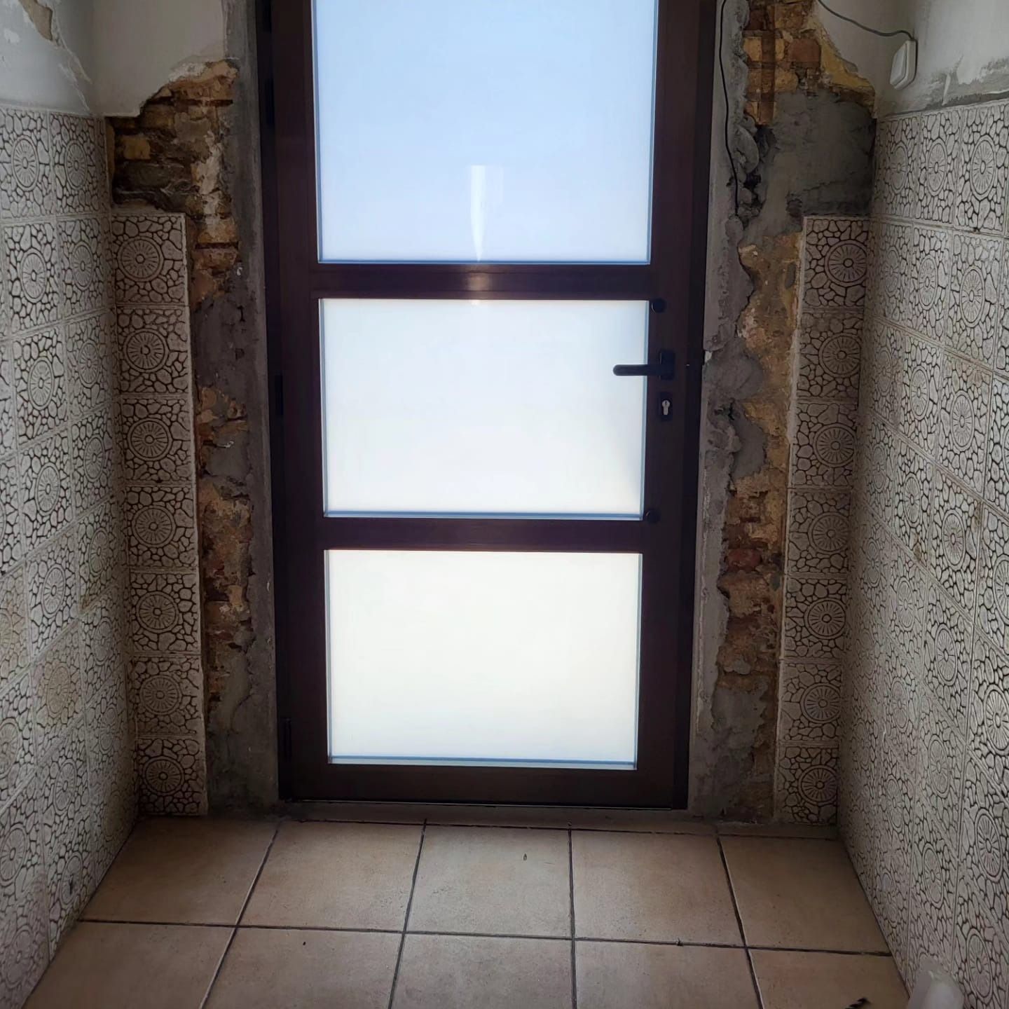 Una puerta de vidrio esmerilado con marco marrón en una habitación pequeña con paredes y piso de azulejos y ladrillos a la vista.