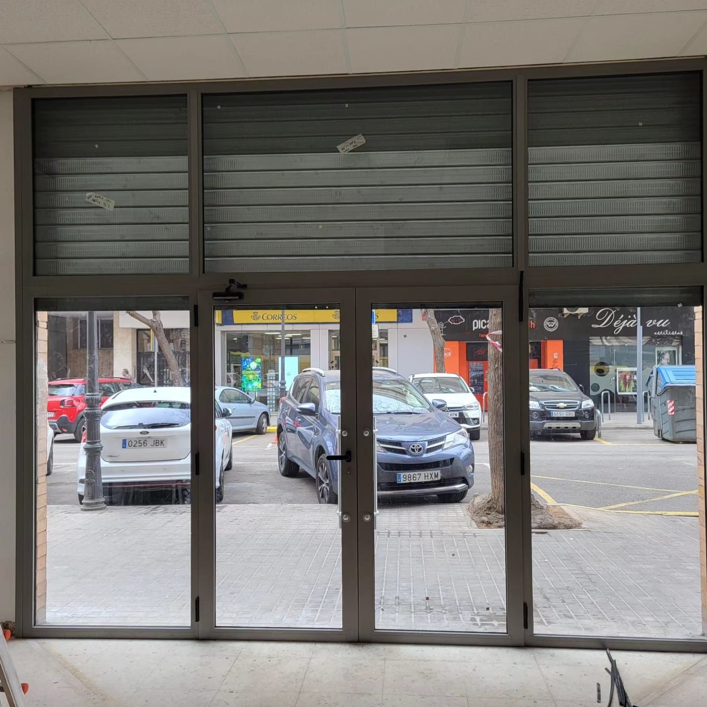 Una tienda con puertas de cristal y una persiana metálica cerrada, vista a la calle. Se ven coches y gente afuera.