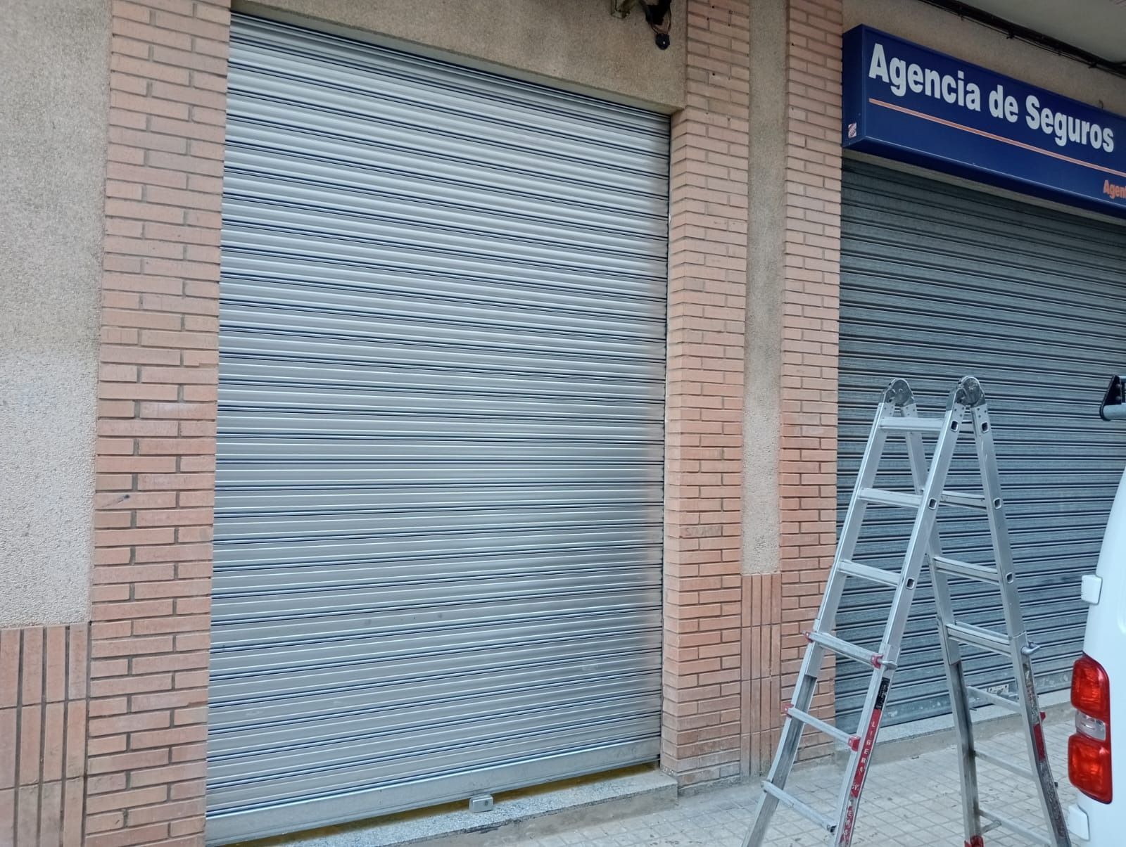 Una persiana metálica de seguridad cerrada cubre la fachada de una tienda. Una escalera se apoya en la pared cerca de un cartel de 