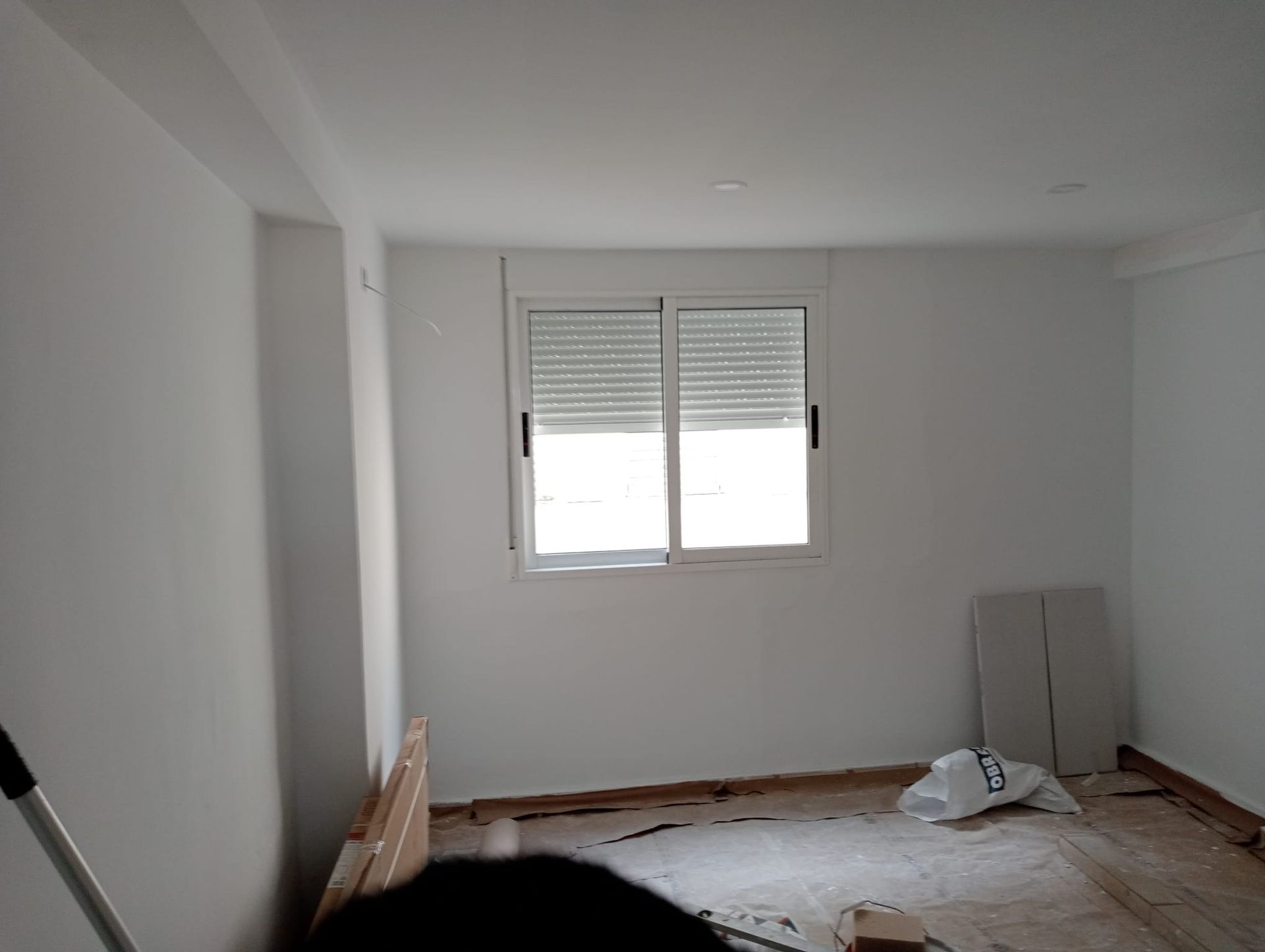 Habitación blanca vacía con ventana, persianas cerradas y materiales dispersos.