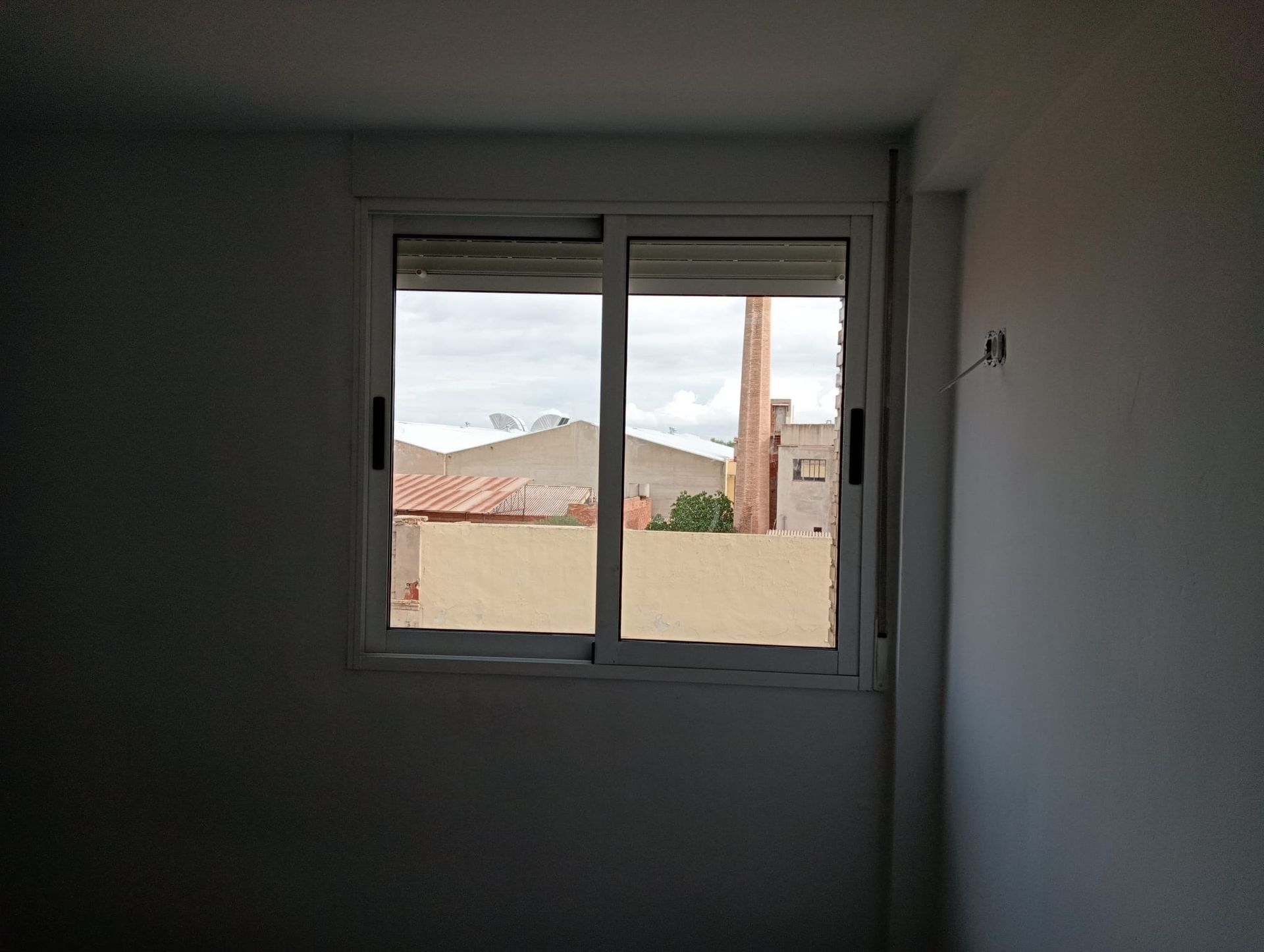 Una ventana con vista a tejados, edificios y una chimenea, en una habitación blanca.