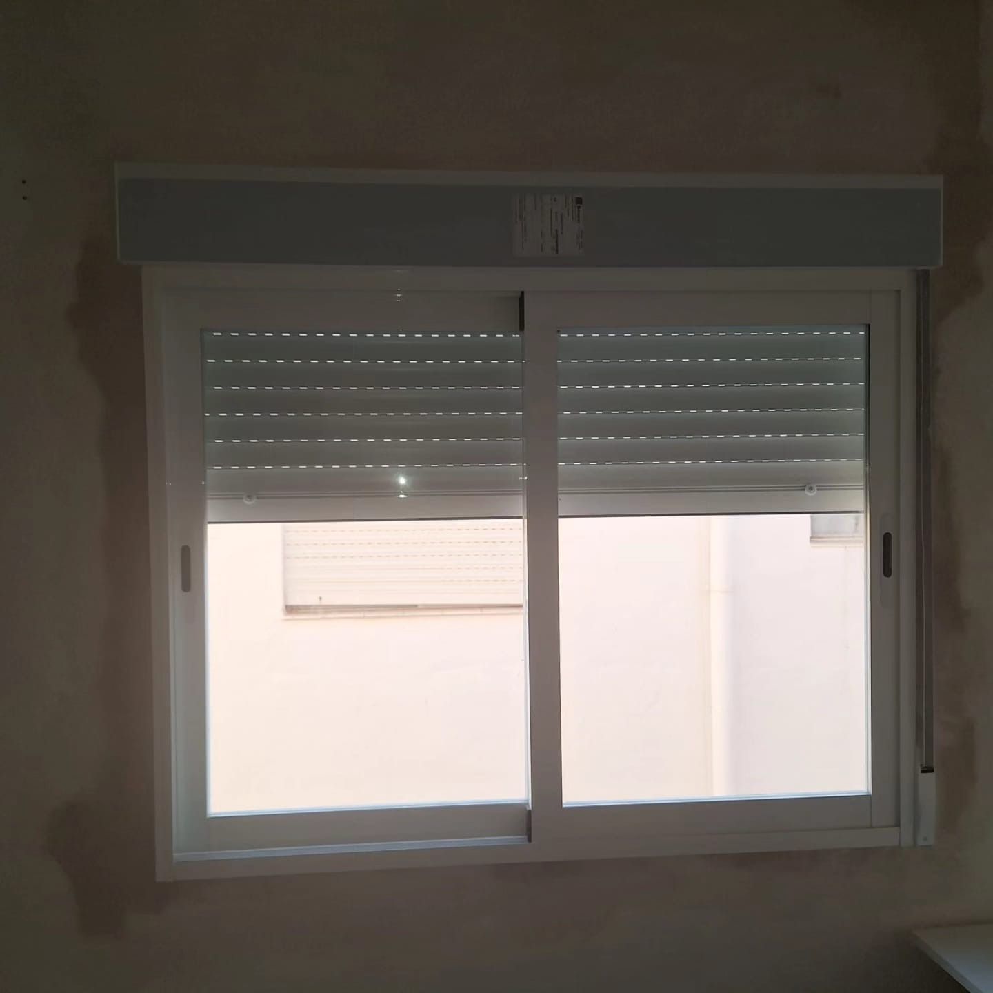 Ventana con marco blanco y persianas grises cerradas. Vista exterior, pared neutra.