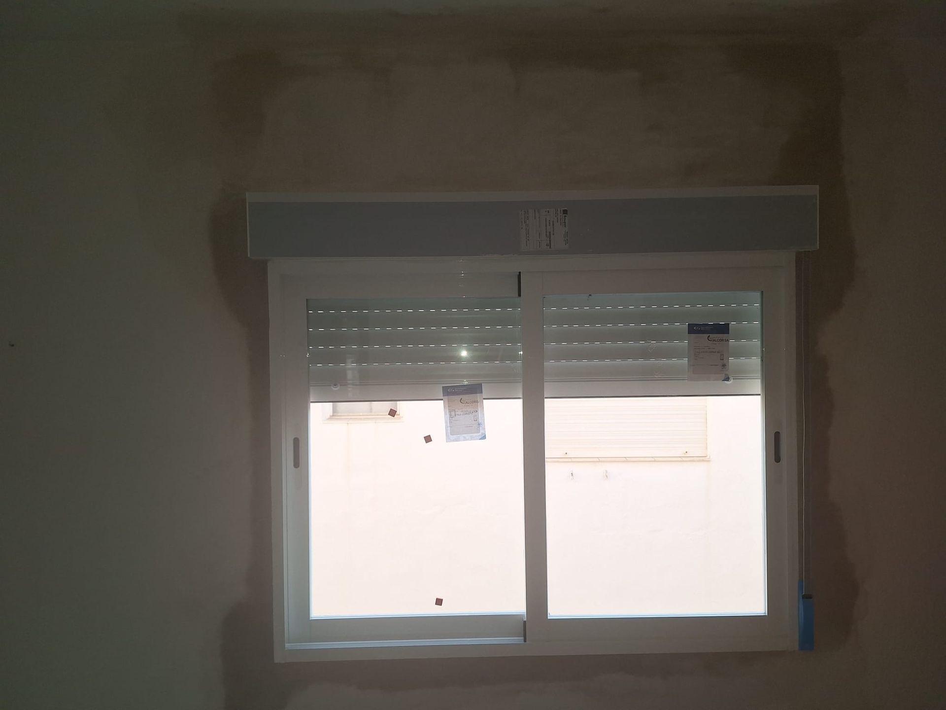 Ventana blanca con persianas cerradas y persiana enrollable sobre pared beige.