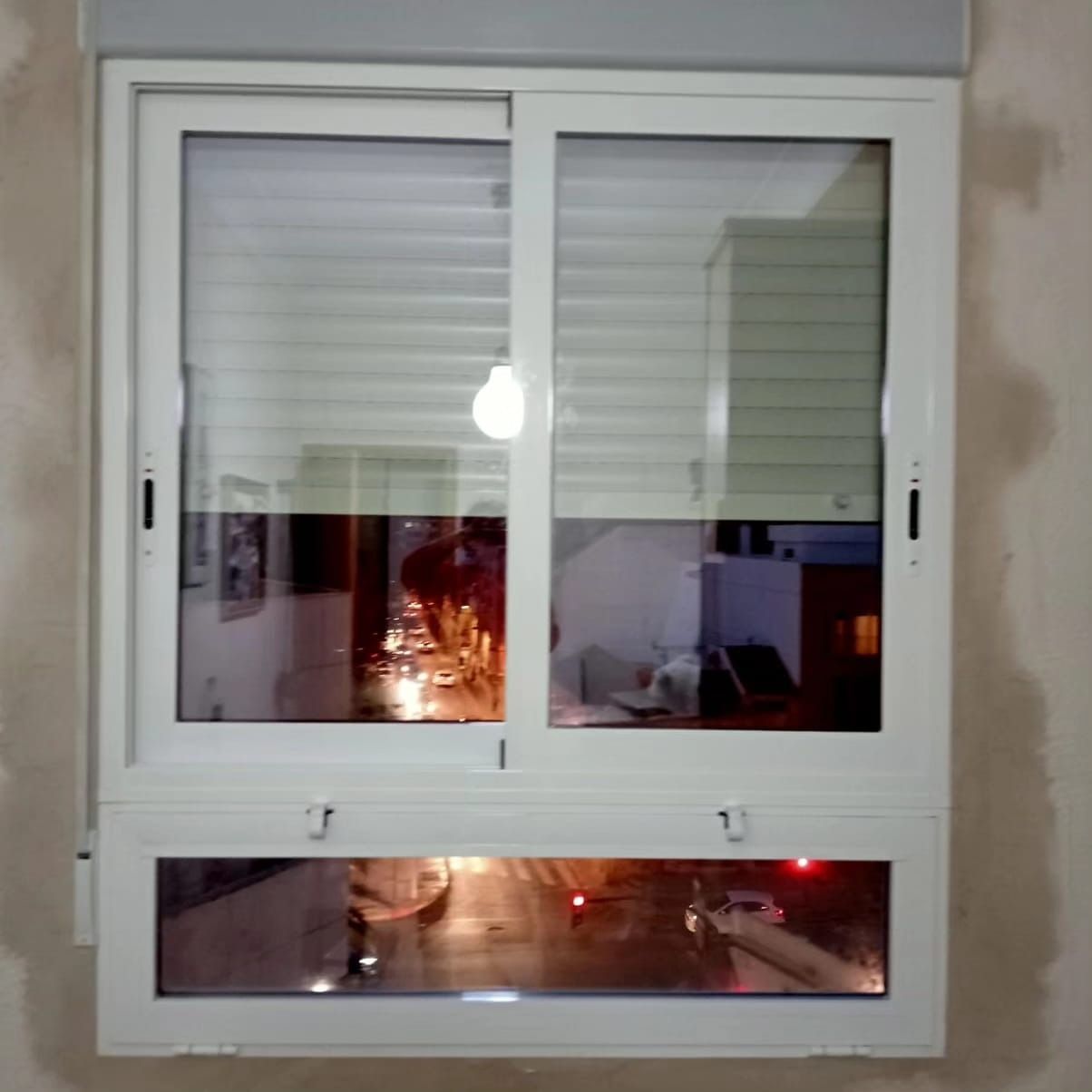 Ventana corrediza blanca con persianas, que refleja una habitación con mucha luz y una ventana inferior.