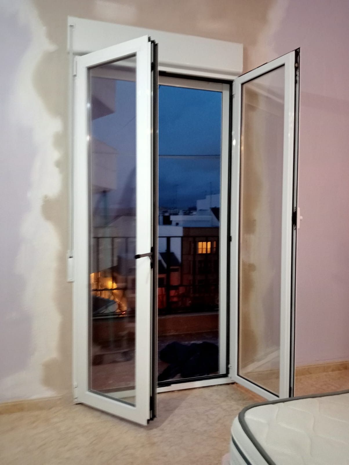 Las puertas dobles blancas se abren a un paisaje urbano nocturno. La luz interior contrasta con la oscuridad exterior.