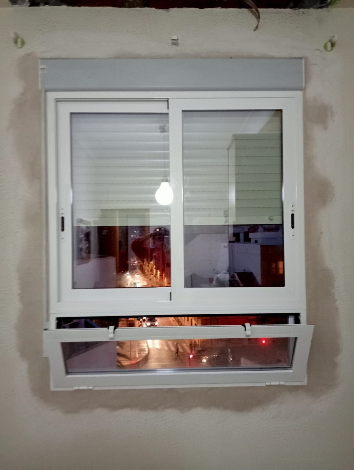 Ventana corrediza blanca con sección inferior abierta, que refleja el interior con persianas blancas.