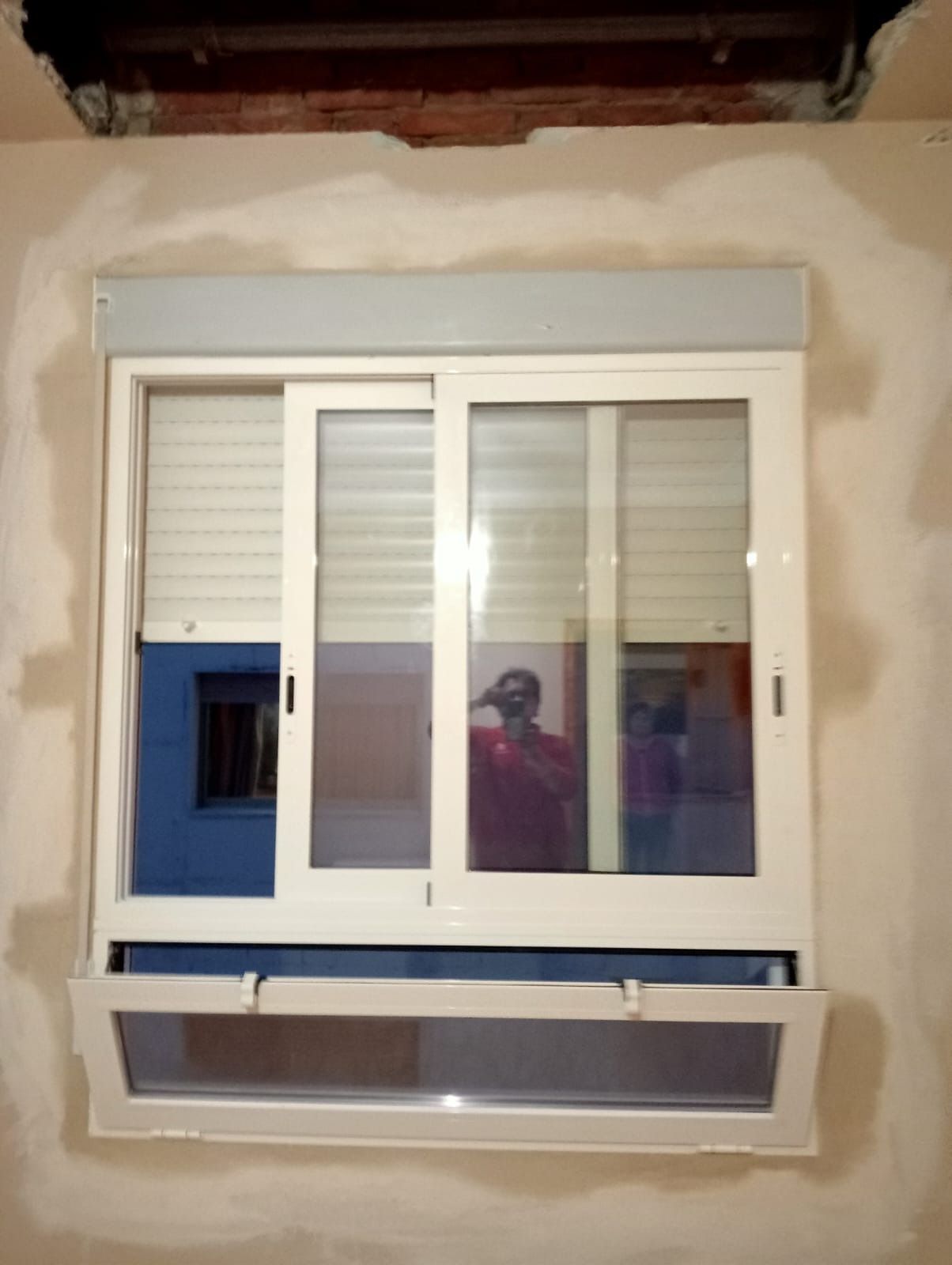 Ventana con marco blanco, persianas y ventana inferior abatible. Una persona se refleja en el cristal.