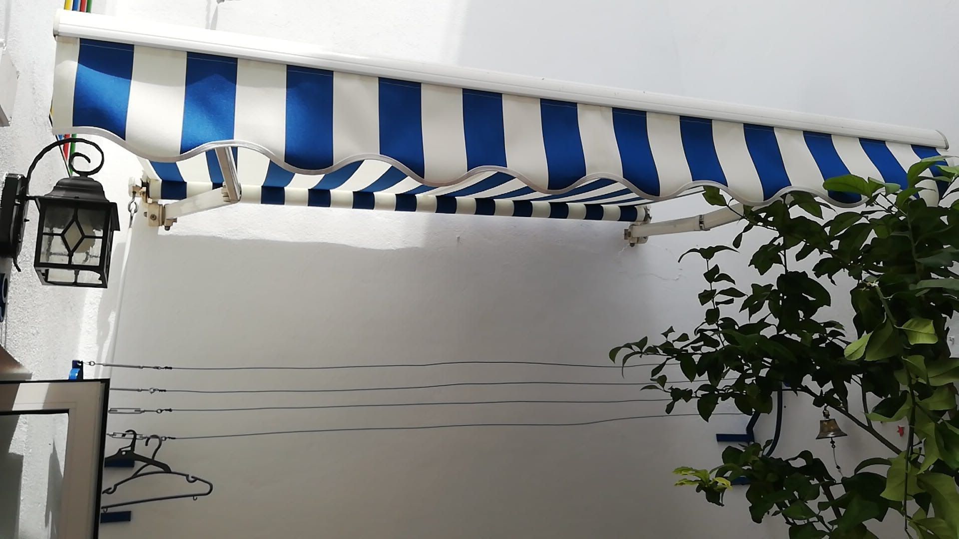 Toldo de rayas azules y blancas sobre un edificio blanco, sobre una planta y un tendedero, con una linterna.
