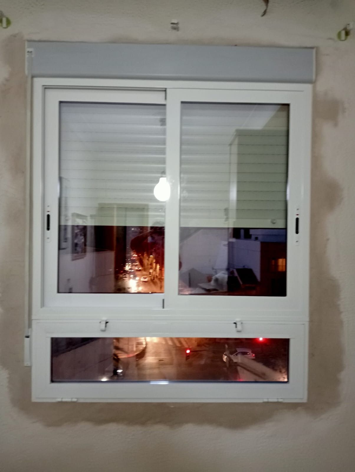Ventana corrediza blanca con persianas y panel inferior que refleja las luces interiores contra una pared neutra.