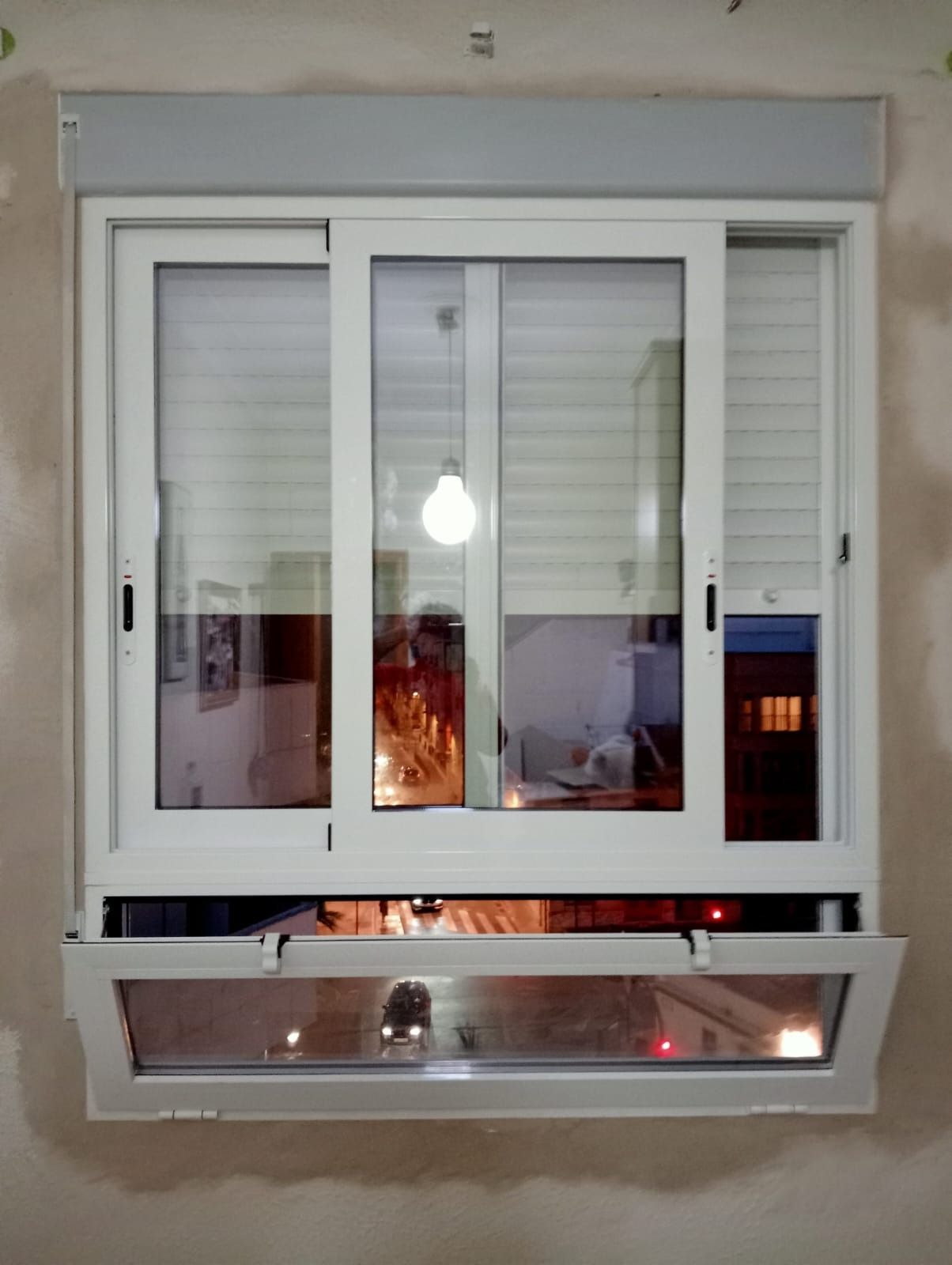 Ventana blanca con persiana retráctil y panel inferior abierto, que refleja una habitación.