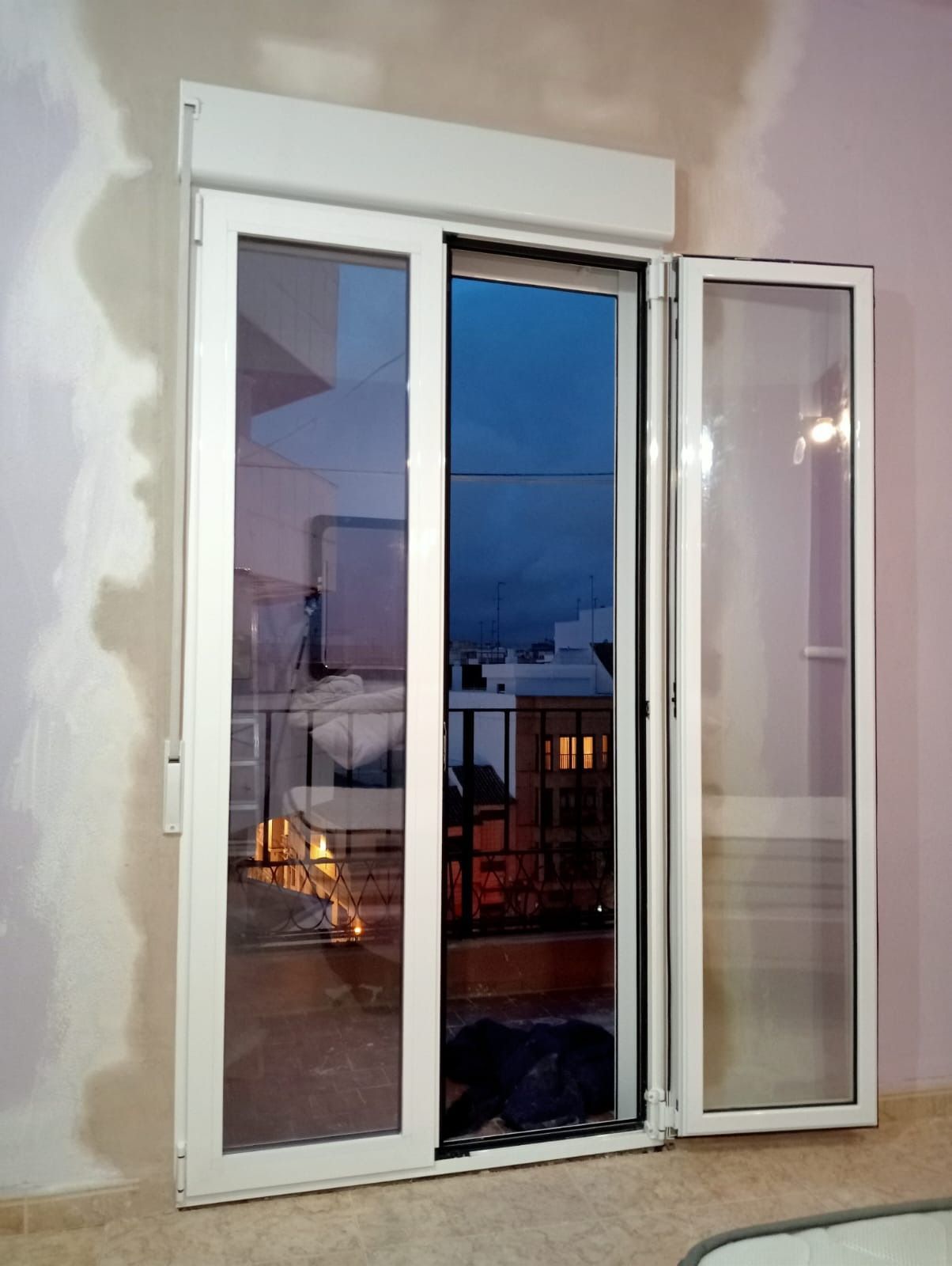 Puerta corrediza de vidrio con marco blanco, parcialmente abierta, que refleja un edificio exterior y un cielo oscuro.