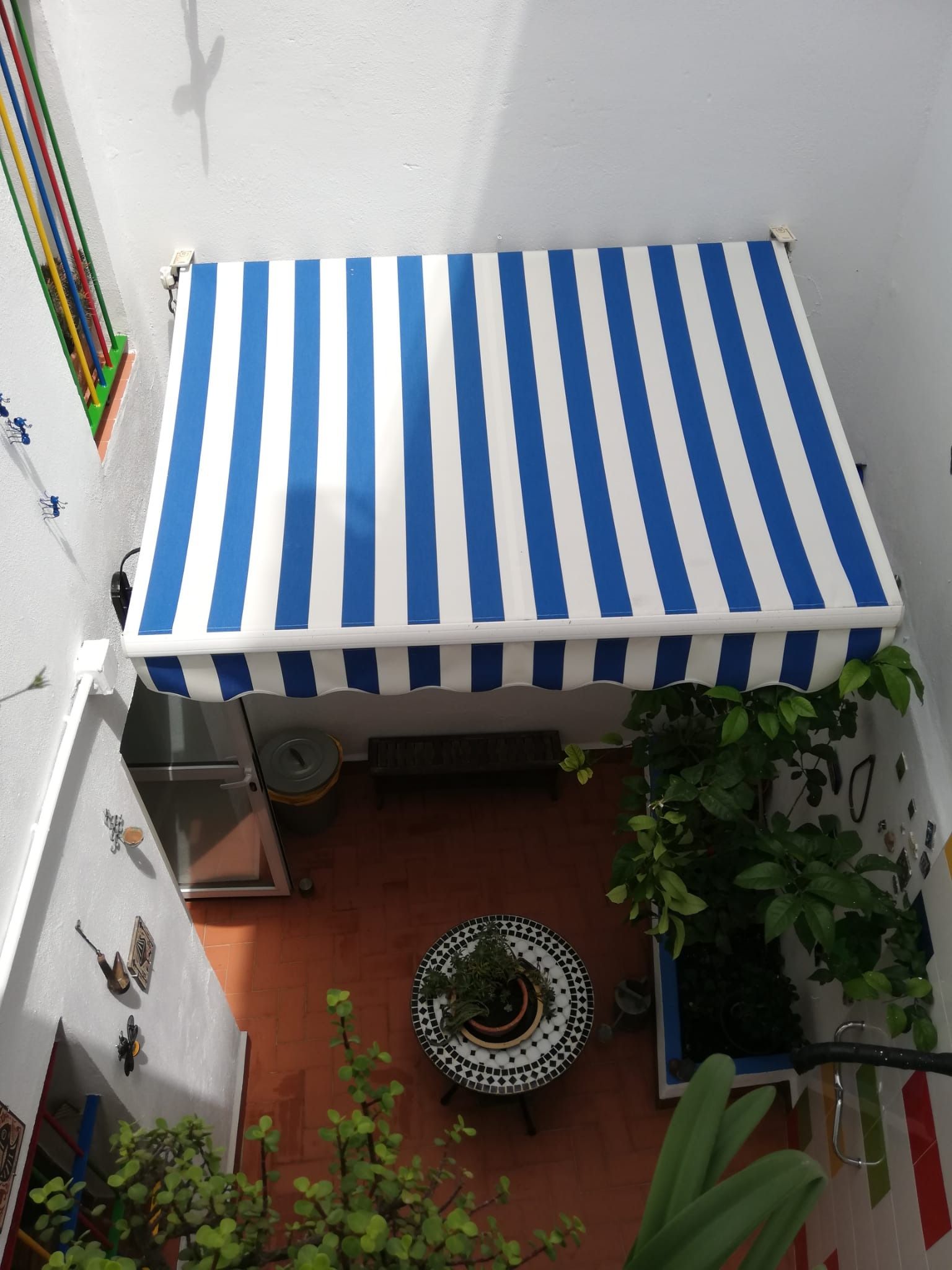 Toldo de rayas azules y blancas que da sombra a un pequeño patio con una mesa y plantas.