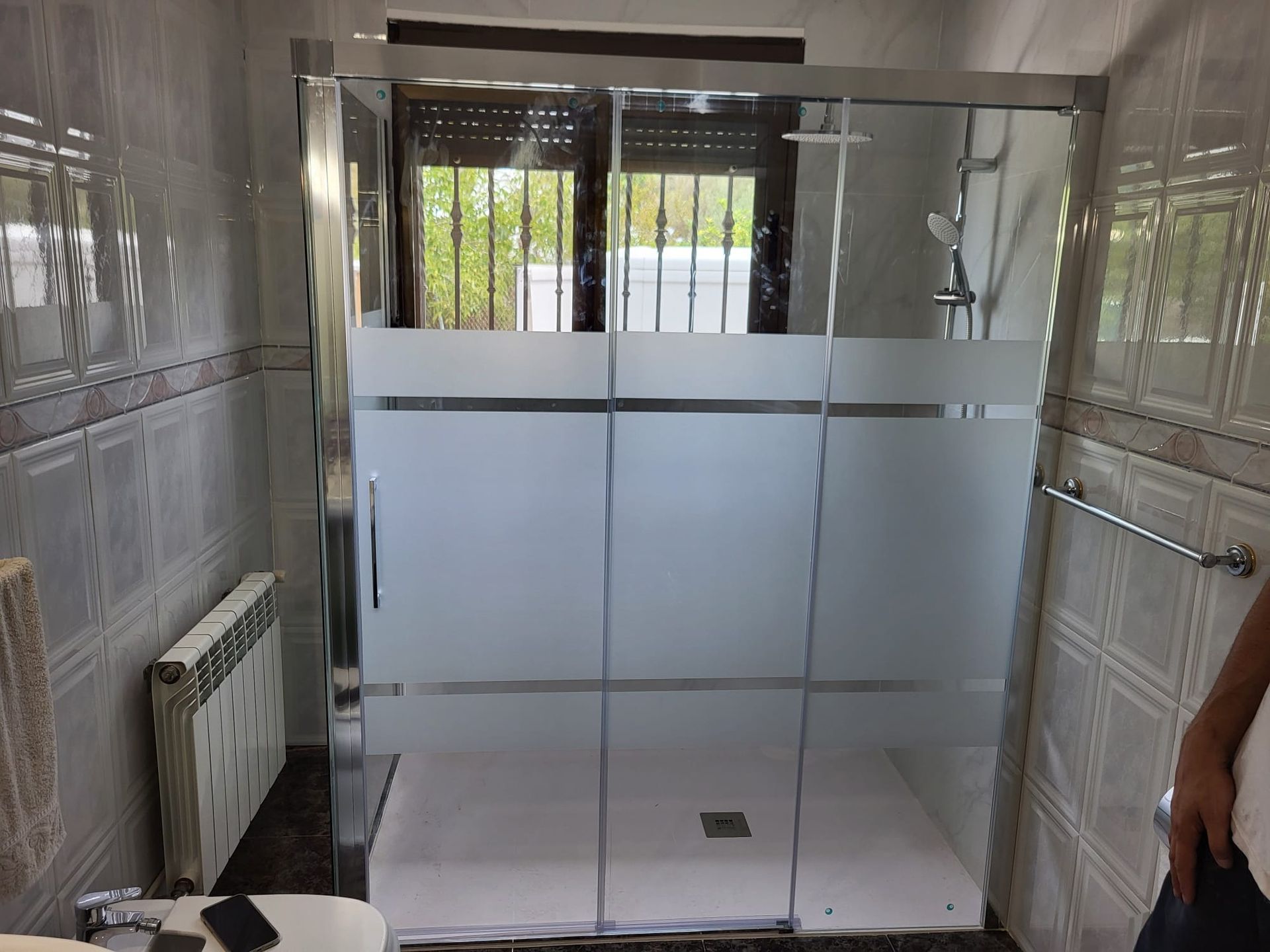 Mampara de ducha en un baño con paneles de vidrio esmerilado. Marco plateado, base de ducha blanca y paredes alicatadas.