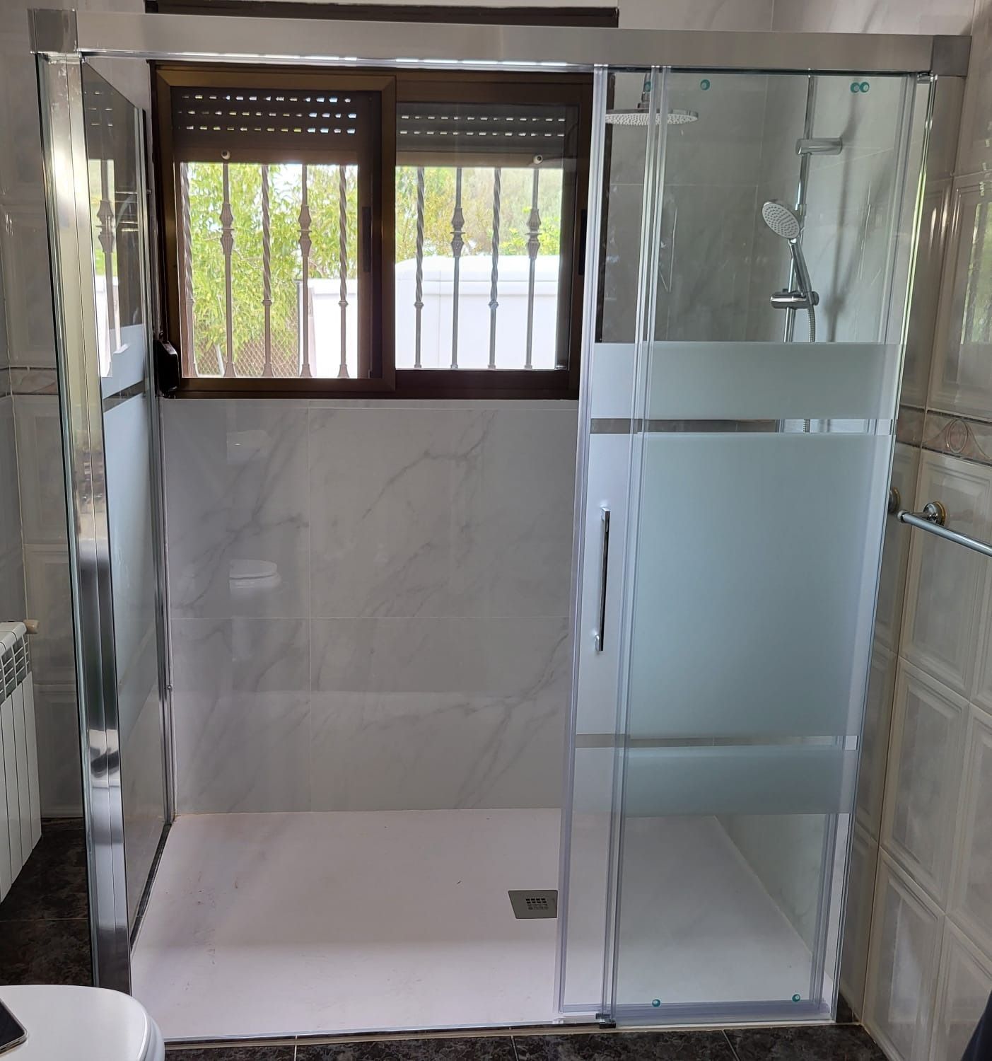 Mampara de ducha con puerta corredera de cristal esmerilado y paredes imitación mármol blanco y ventana.