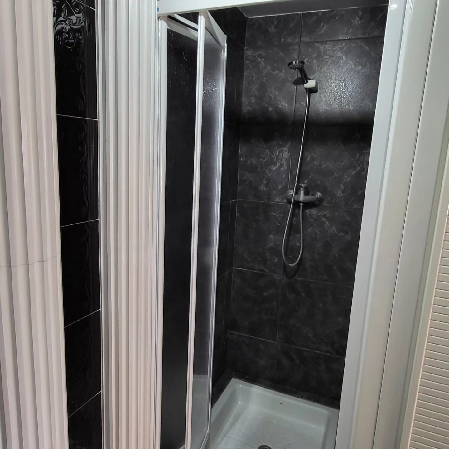 Cabina de ducha con paredes de azulejos oscuros, marco plateado y cortina blanca.