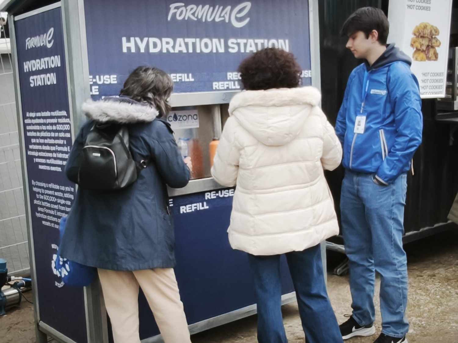 Tres personas en un puesto de hidratación de Fórmula E, llenando vasos en un punto de bebidas azul.