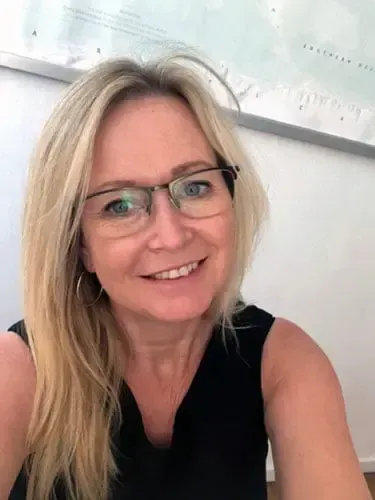 Mujer rubia sonriente con gafas y una blusa negra sin mangas, sentada en el interior contra una pared blanca.