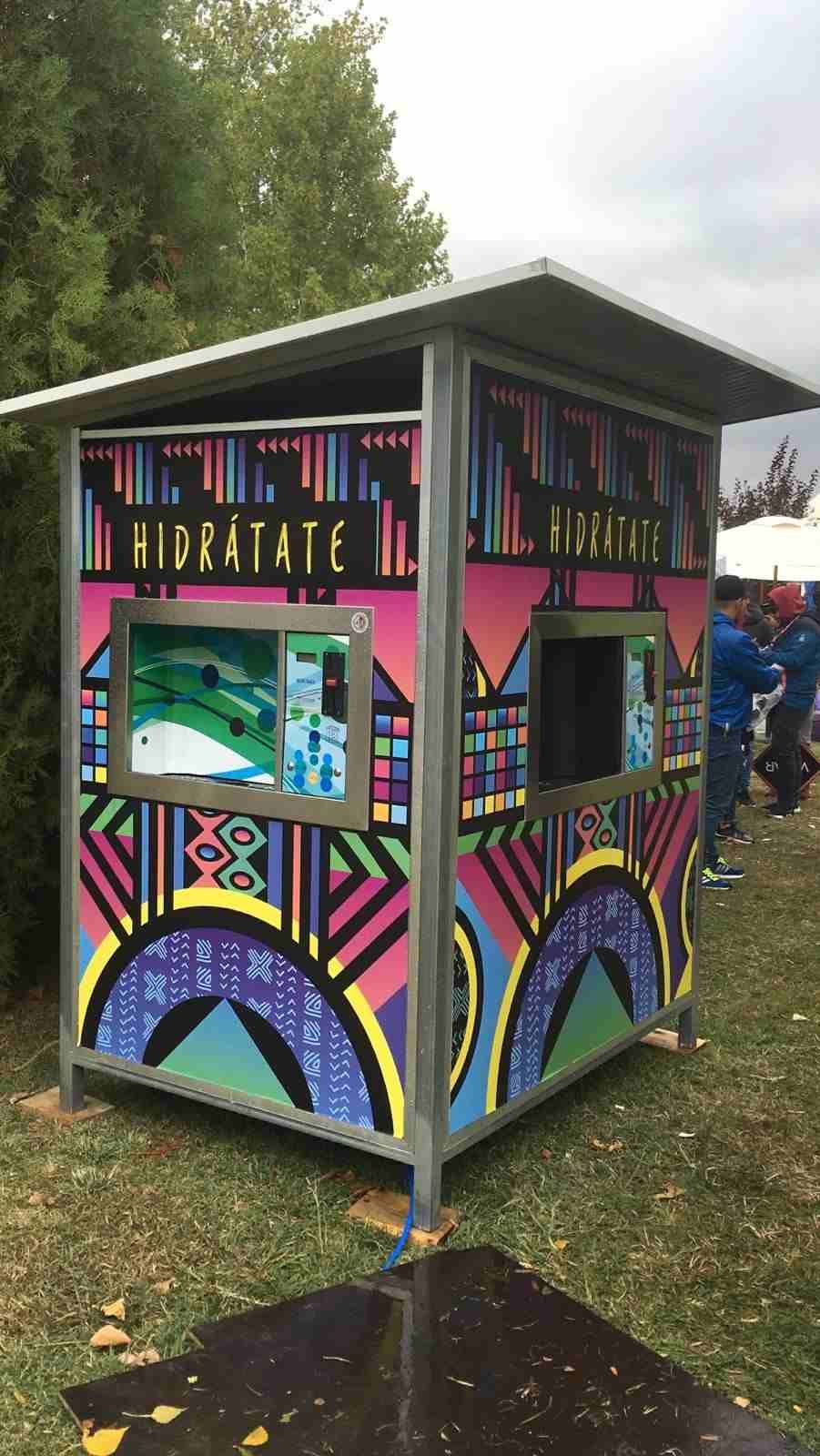 Pequeño quiosco blanco con techo y ventana azules, situado al aire libre junto a un muro de hormigón y una ladera verde.