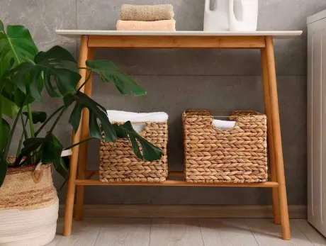 Estante de madera con dos cestas tejidas, toallas apiladas y una botella de detergente. A la izquierda hay una maceta con una planta.