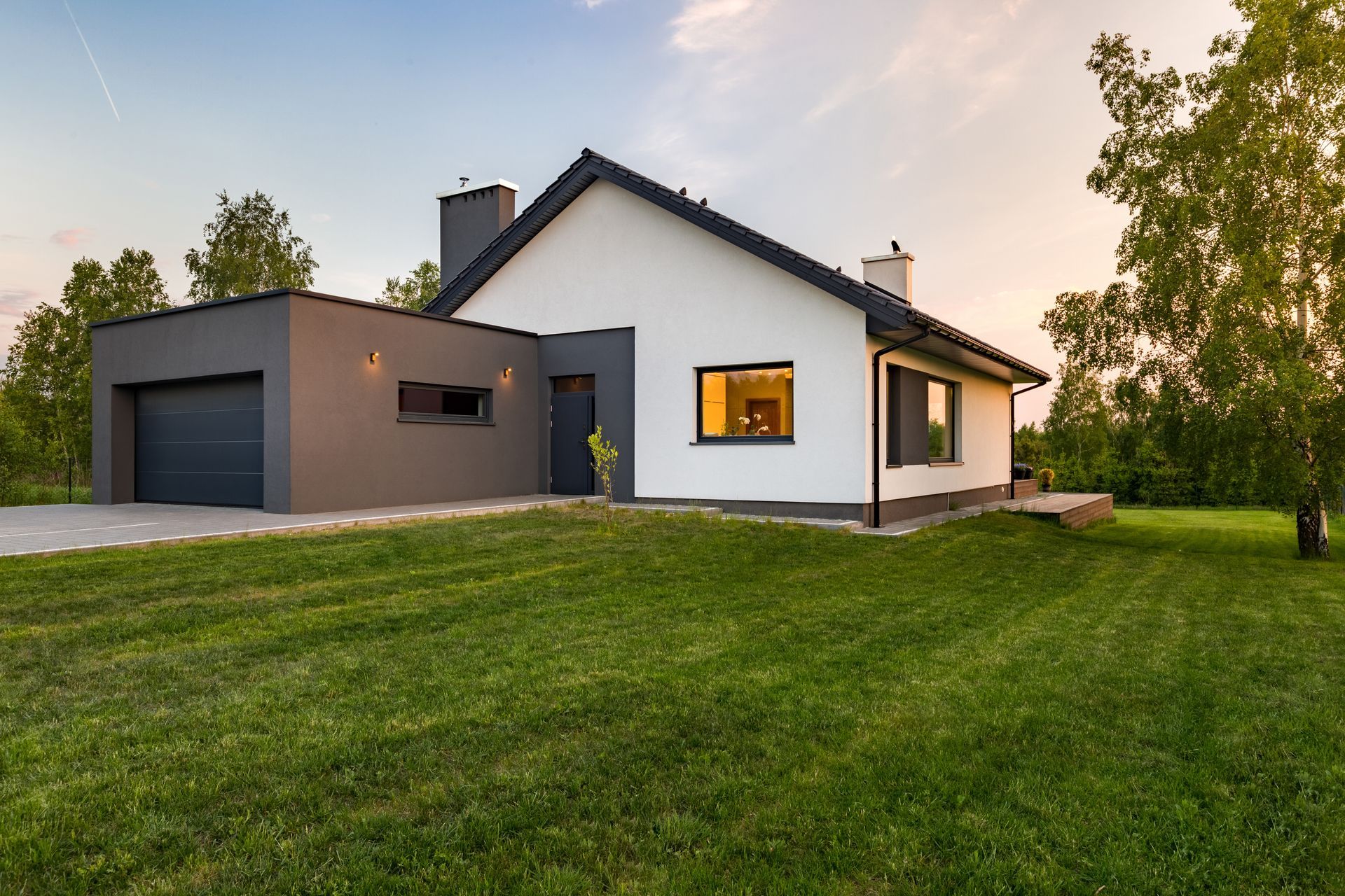 Maison moderne blanche et grise avec un garage, des fenêtres éclairées et une grande pelouse verte au coucher du soleil.