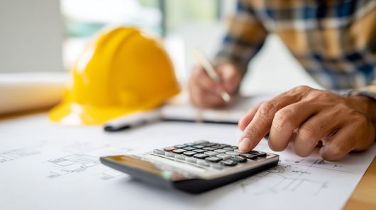 Un homme en train d’utiliser une calculatrice sur un plan, avec un casque de chantier jaune en arrière-plan.