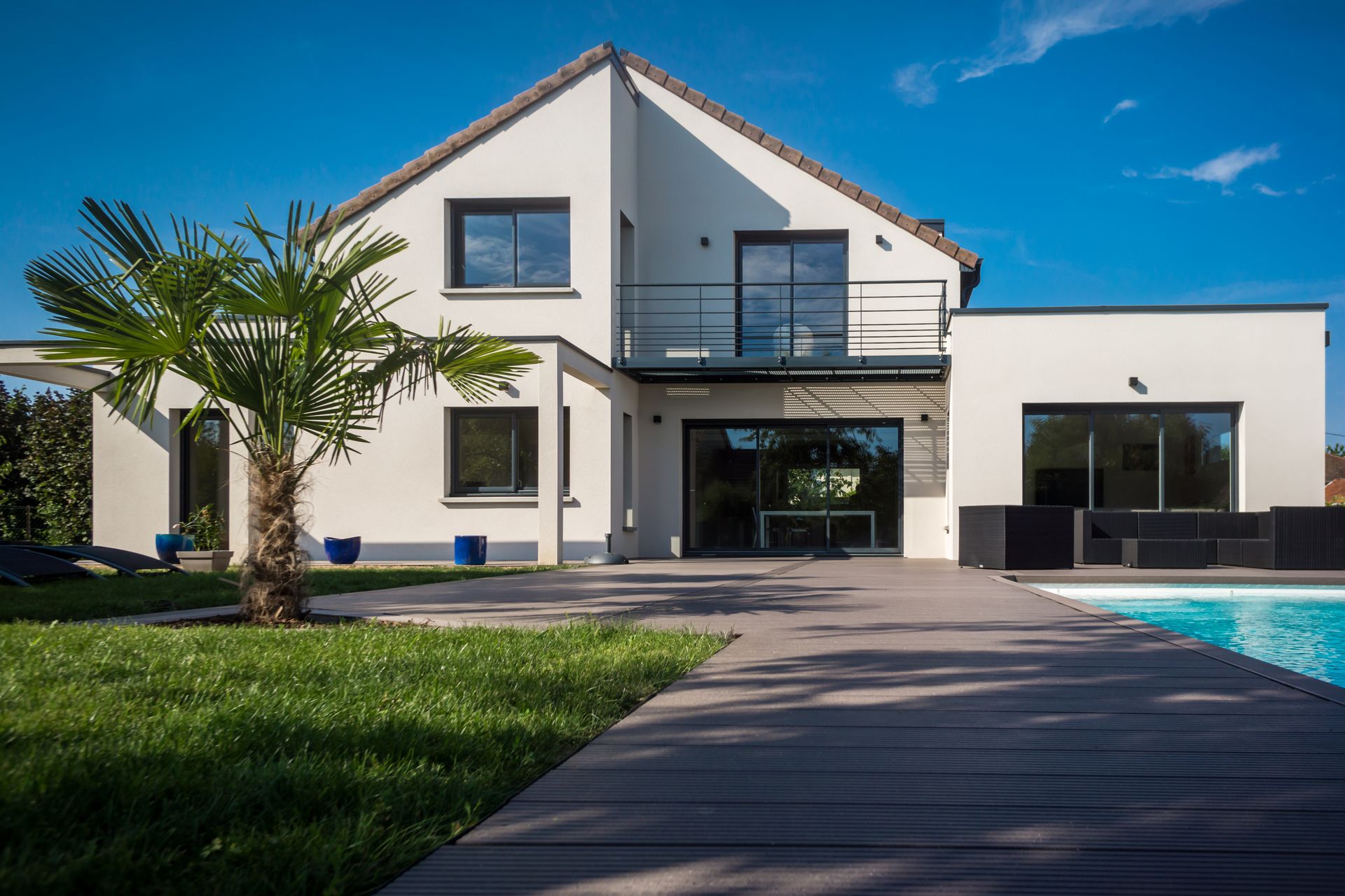 Maison moderne blanche à deux étages, avec une piscine, une terrasse et un palmier sous un ciel bleu.