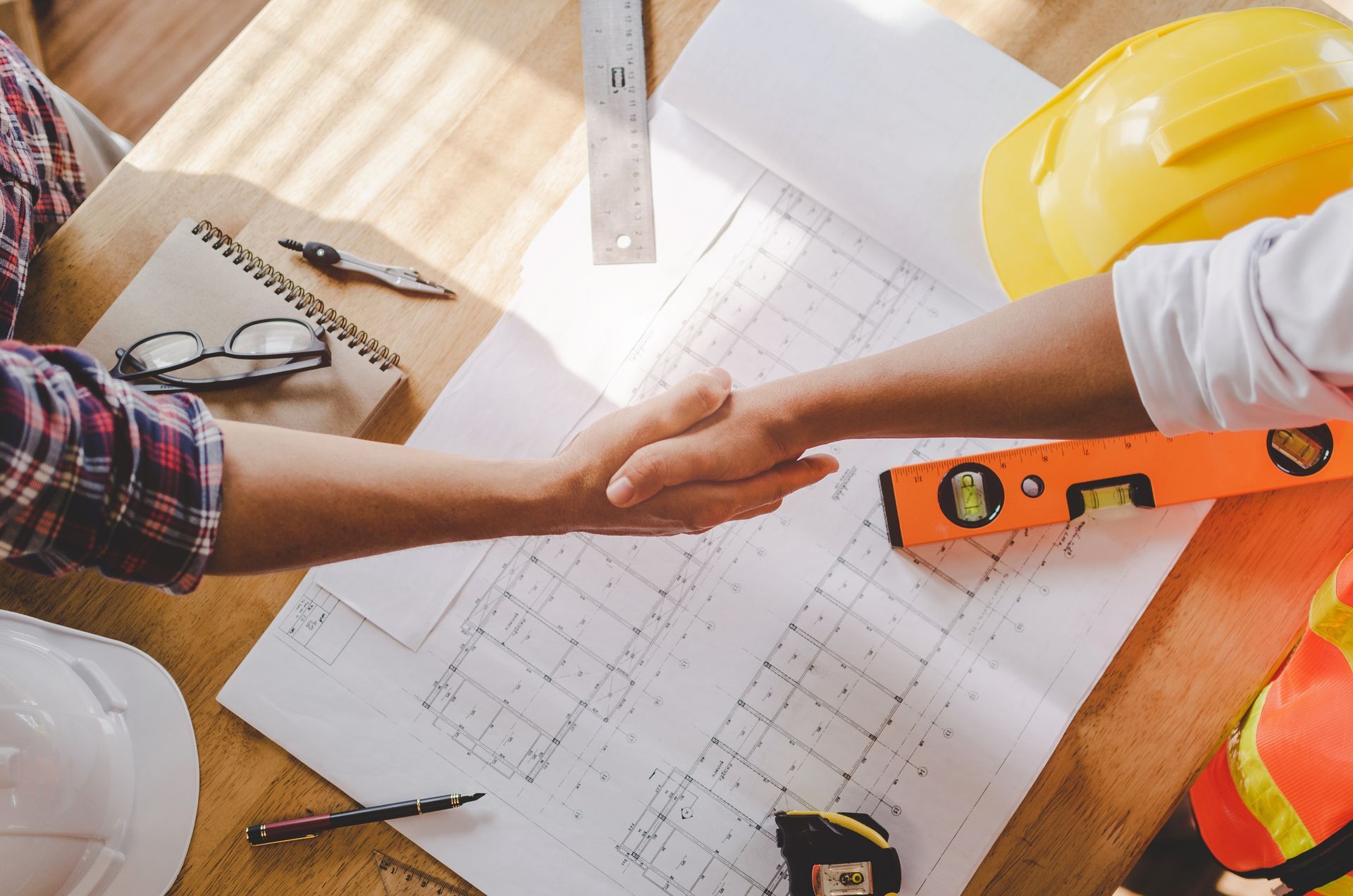 Deux ouvriers se serrent la main au-dessus de plans, avec un casque de chantier, une règle et des plans de construction sur un bureau.
