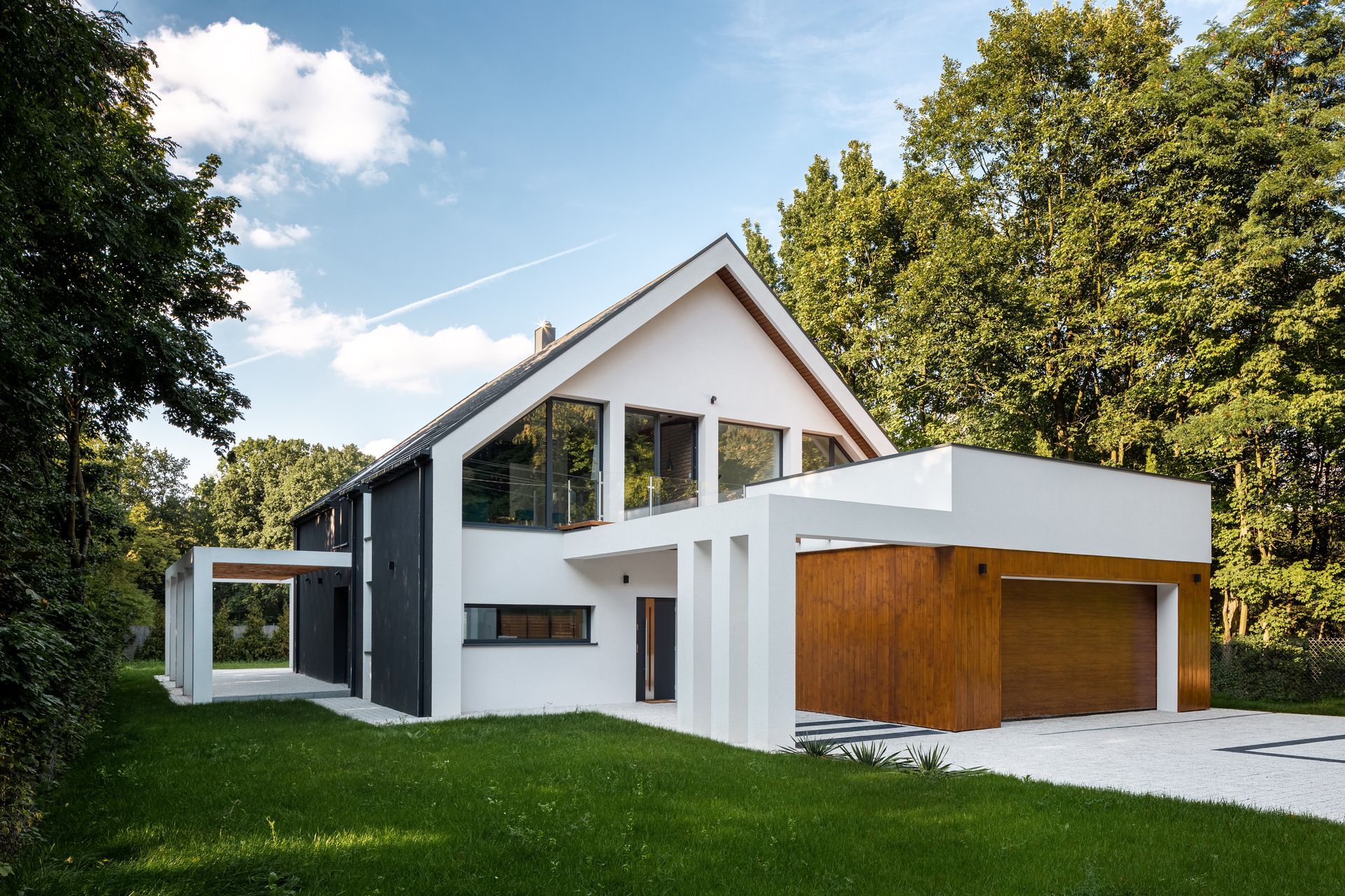 Maison moderne blanche avec un toit pentu, une porte de garage en bois et une pelouse verdoyante sous un ciel bleu.