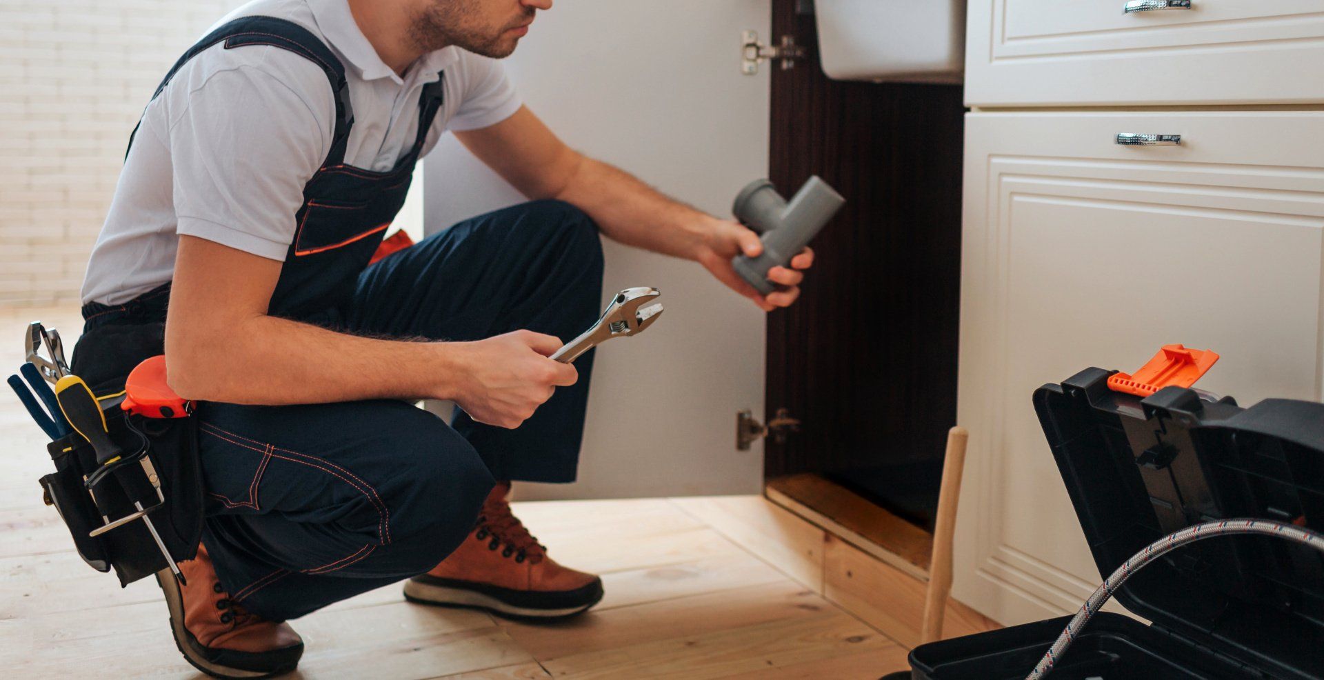 Un technicien agenouillé à côté d'une armoire, utilisant une clé plate et une sacoche à outils lors d'une réparation à domicile.