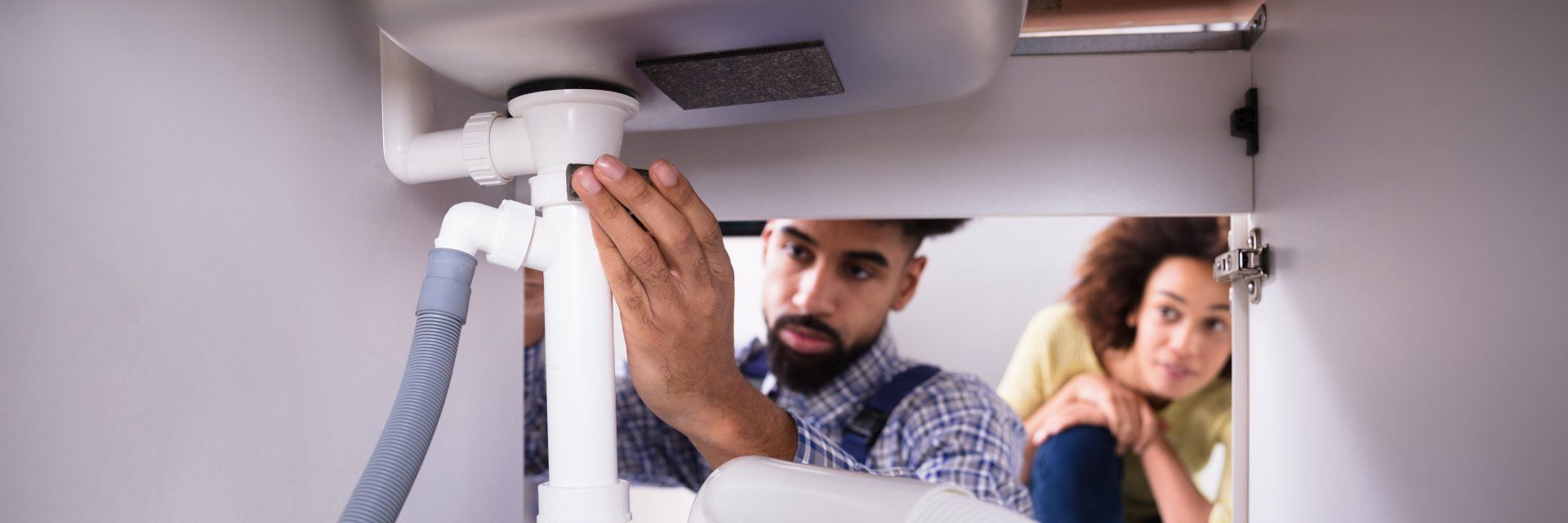 Un homme installe un tuyau d'évier sous un meuble, sous le regard d'une autre personne à proximité.