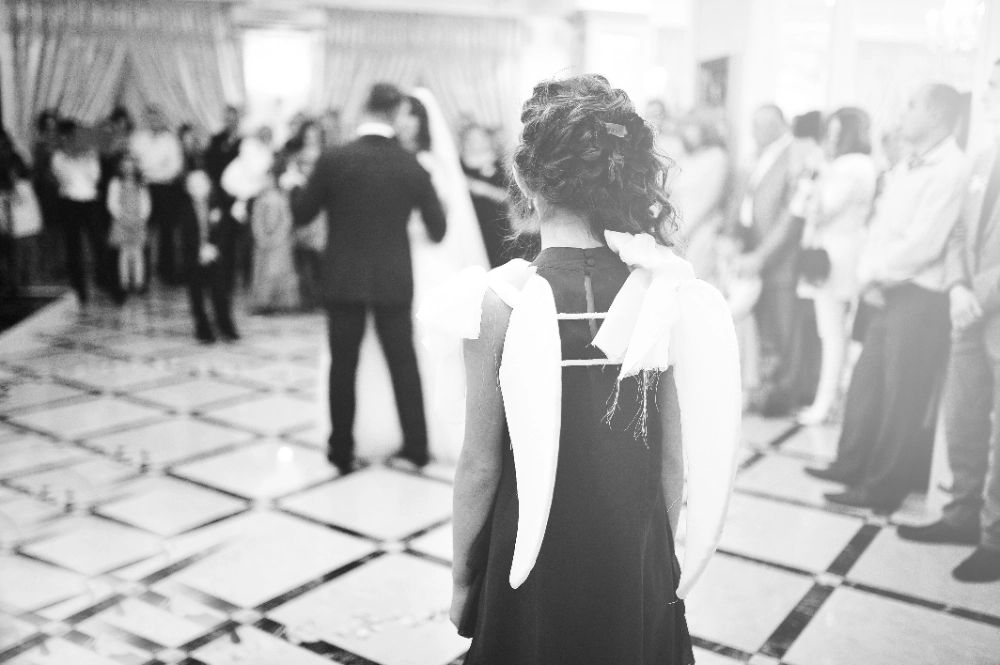 Uma menina com asas de anjo está parada na frente dos noivos em uma recepção de casamento.