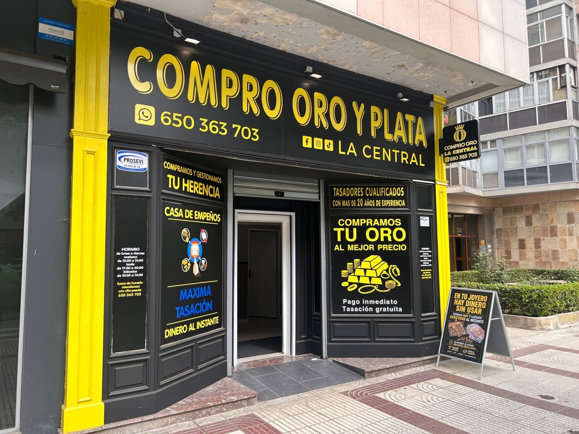 Fachada de «COMPRO ORO Y PLATA» con letreros negros y amarillos y un cartel tipo caballete en la acera
