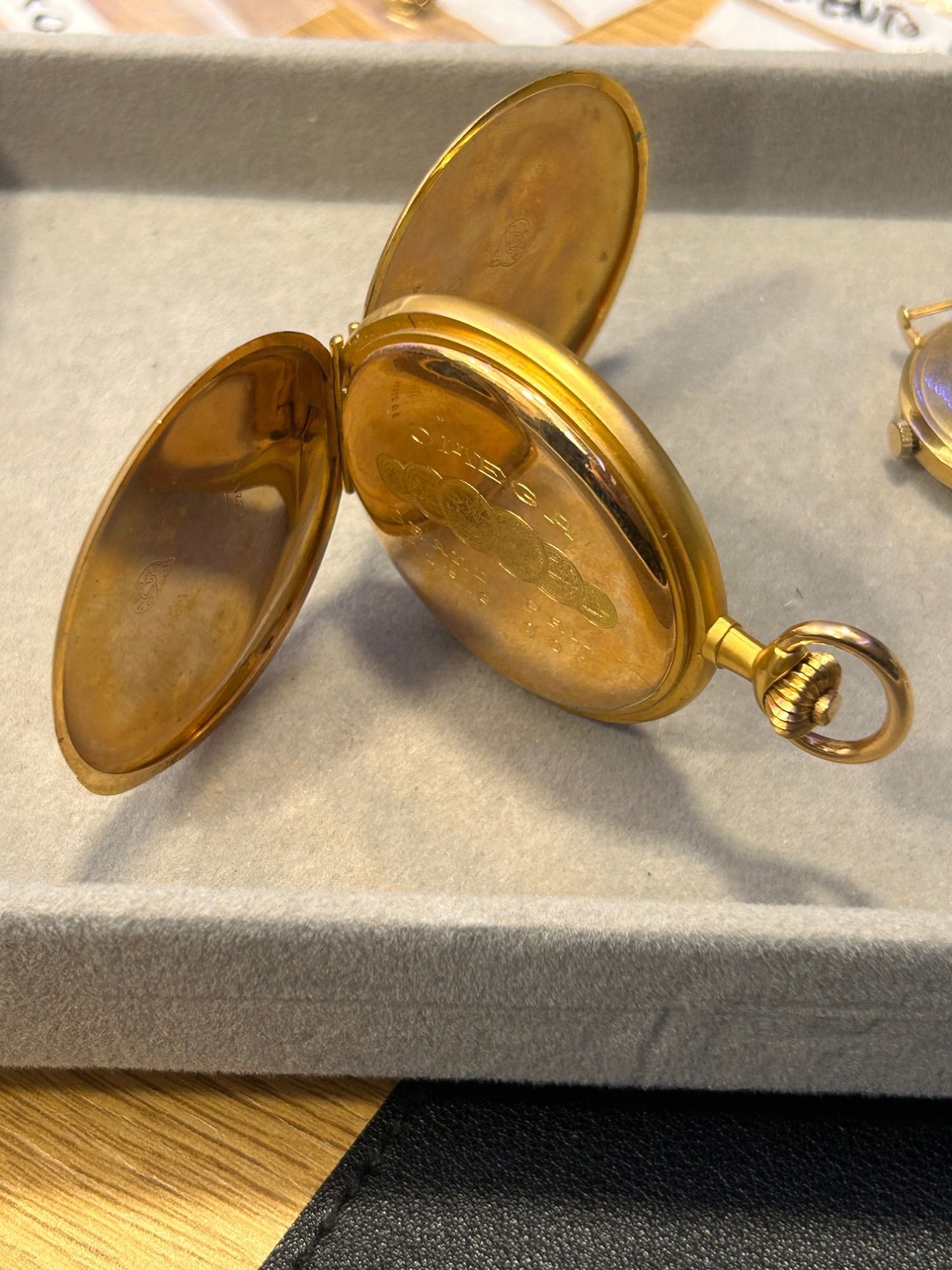 Reloj de bolsillo dorado con la tapa abierta sobre una bandeja de exhibición gris.