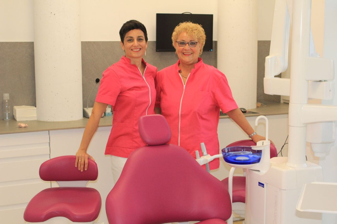 Dos mujeres están de pie junto a una silla dental en un consultorio dental.