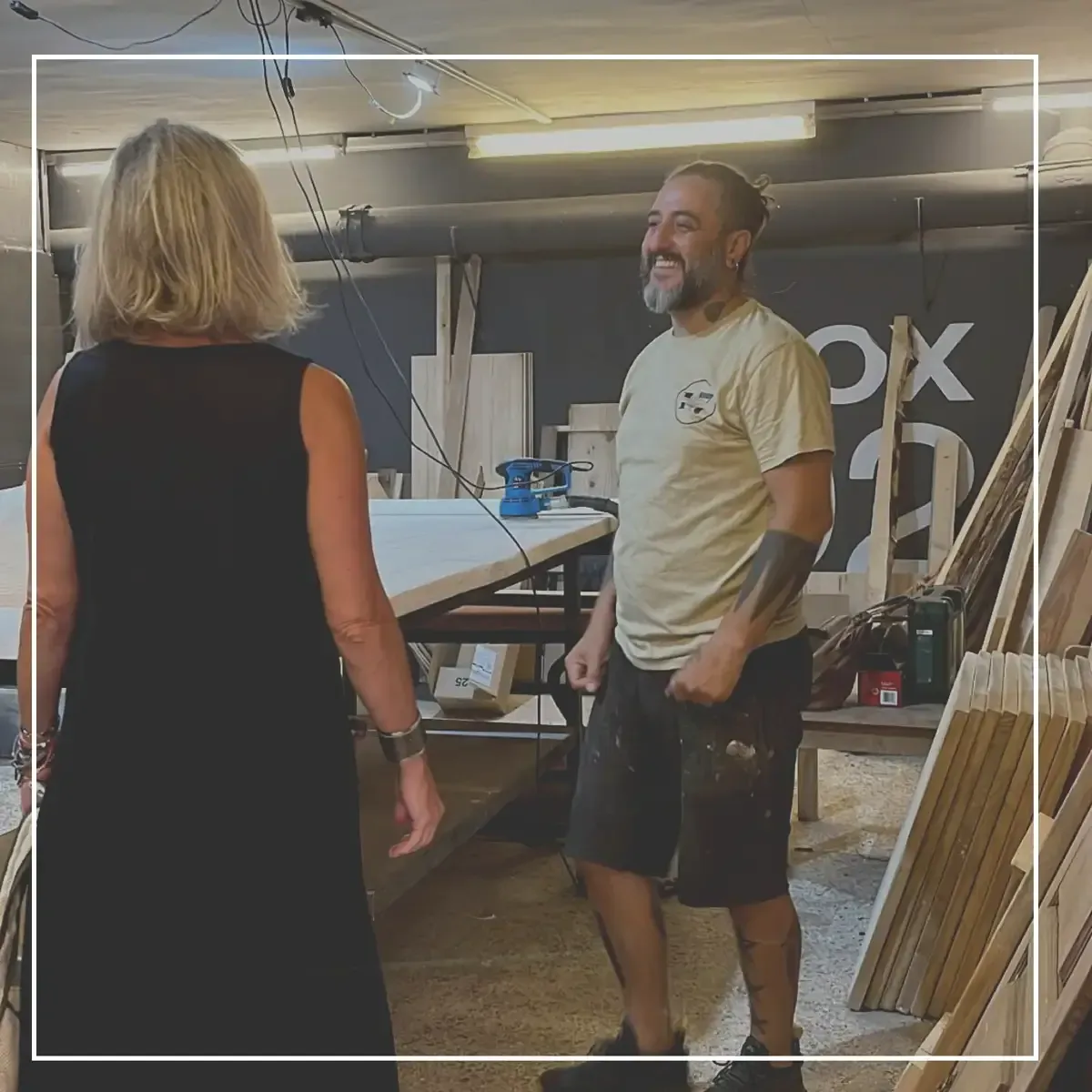 Dos personas conversando en un taller, con una mesa de trabajo, herramientas y tablas apiladas al fondo.