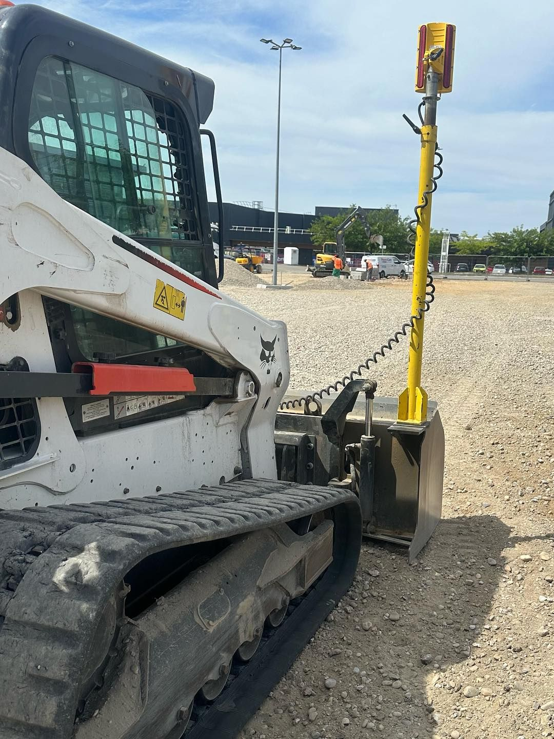 Chargeuse compacte jaune avec attelage à chaîne sur un terrain en gravier, vue de côté.