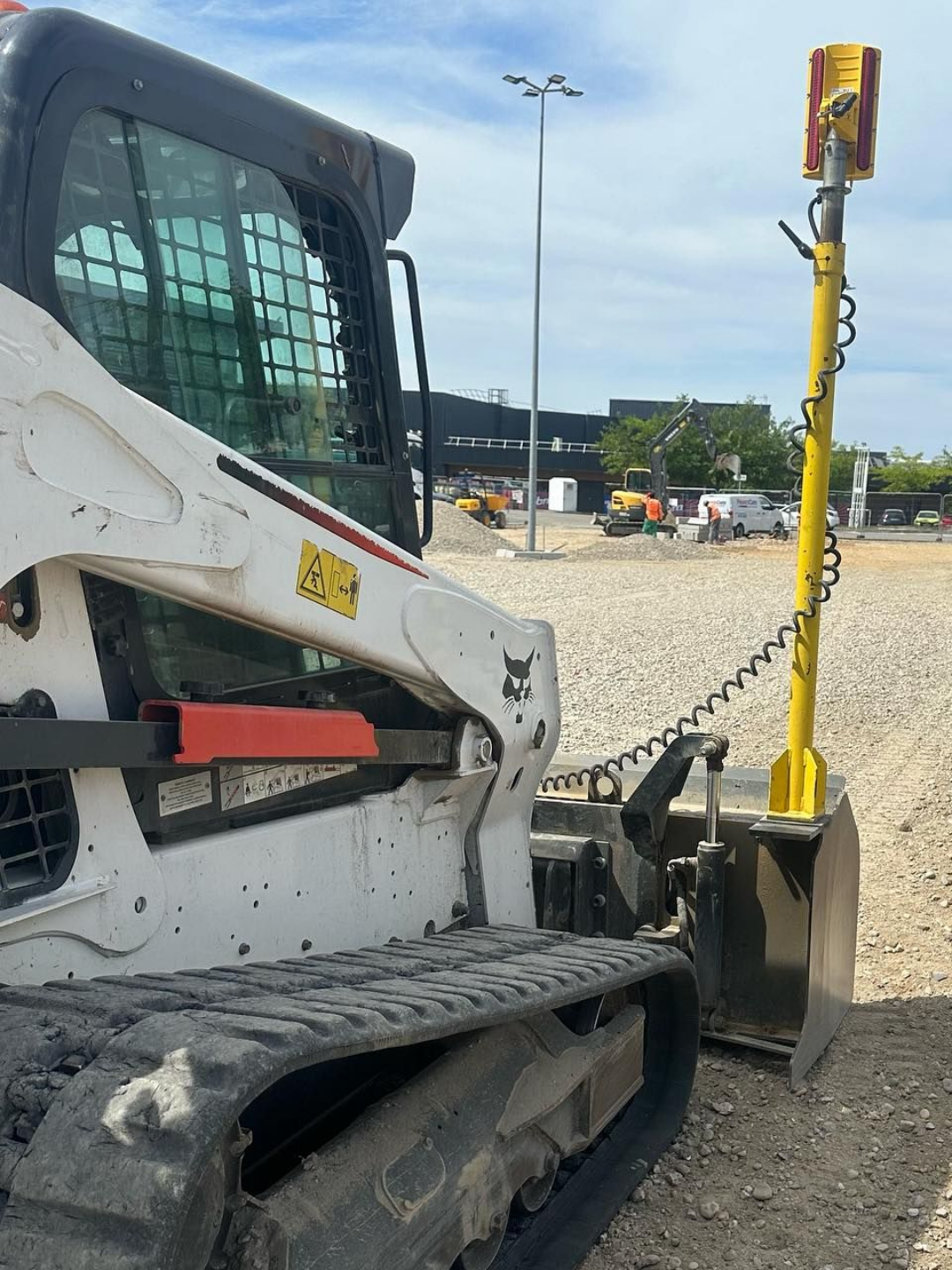 Chargeuse compacte blanche à chenilles avec accessoire jaune sur du gravier dans un chantier.